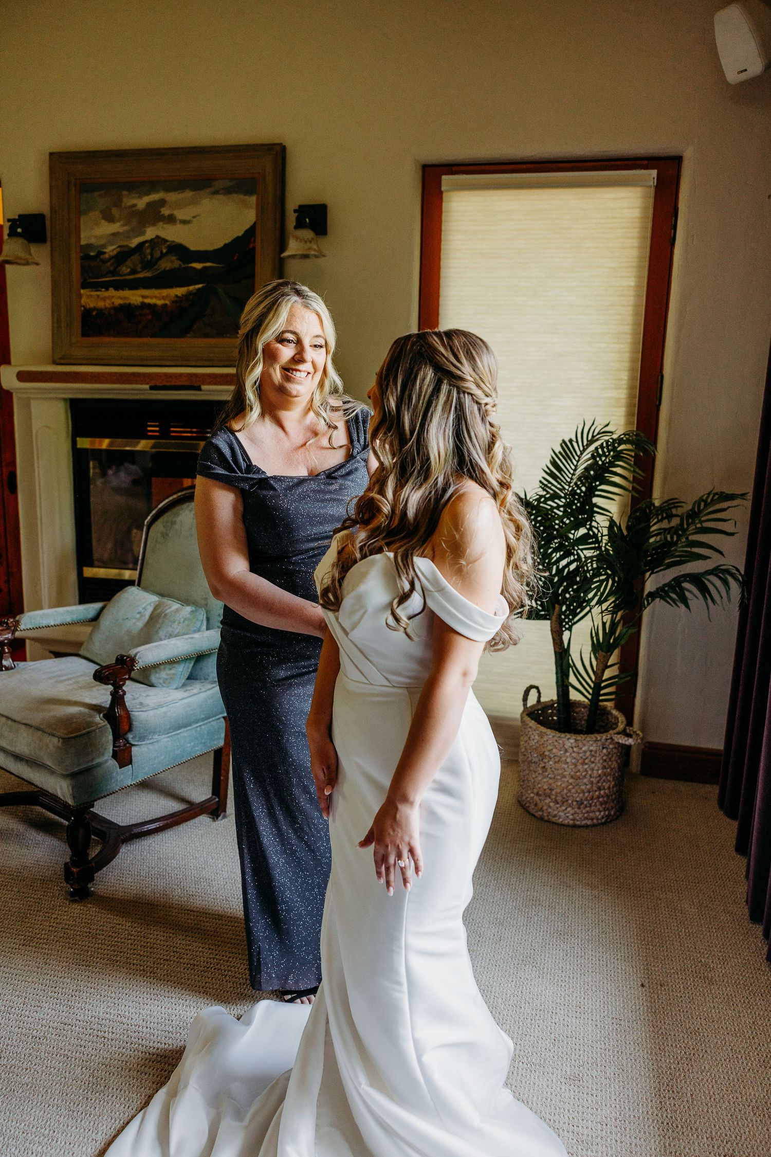Two people share a joyful moment as one adjusts the other's elegant white wedding dress in an indoor setting.