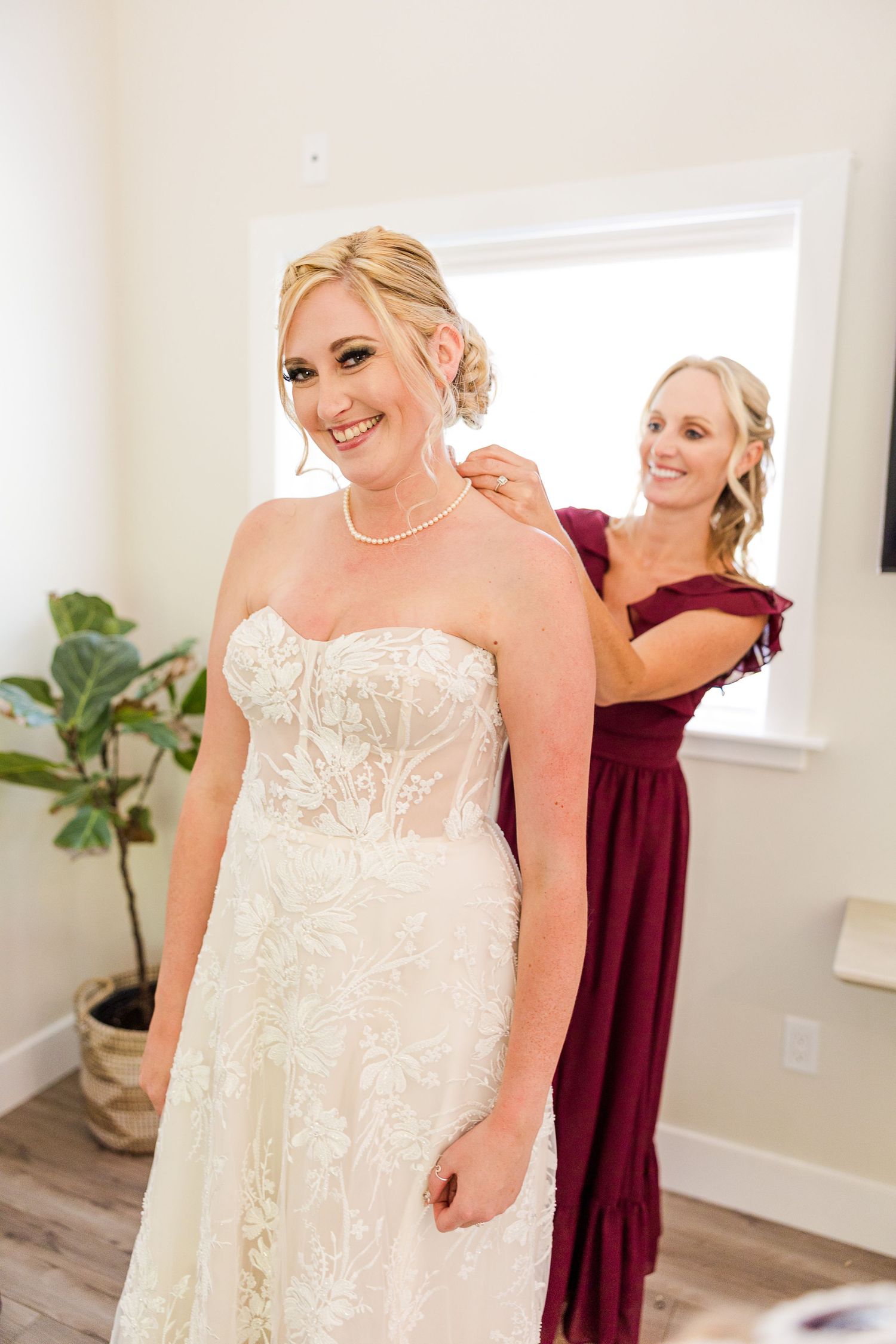 Bride in strapless lace gown stands elegantly while bridesmaid in burgundy helps with final touches.