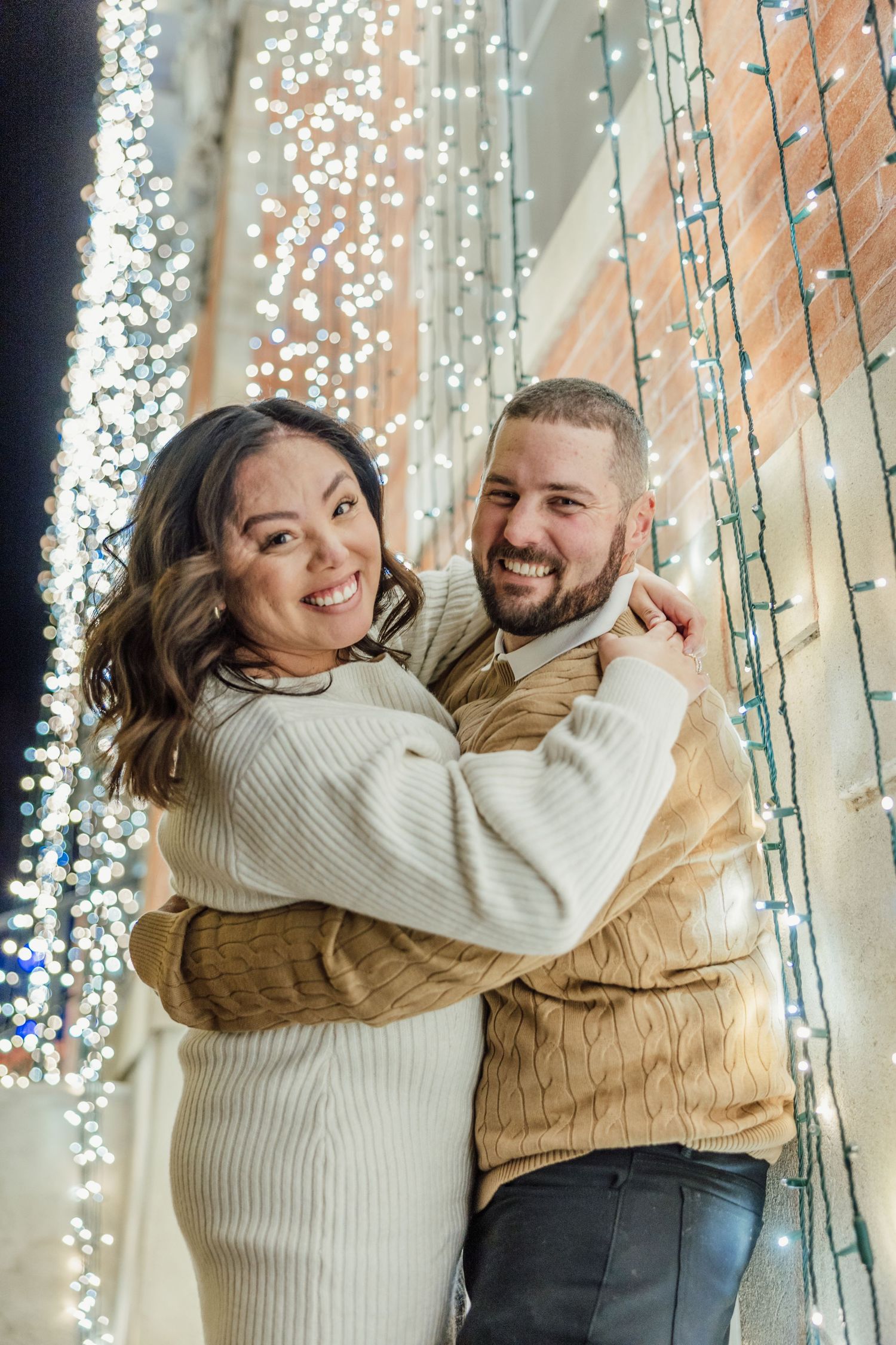 A couple laughs and embraces while posing against a backdrop of twinkling string lights in a festive holiday setting.