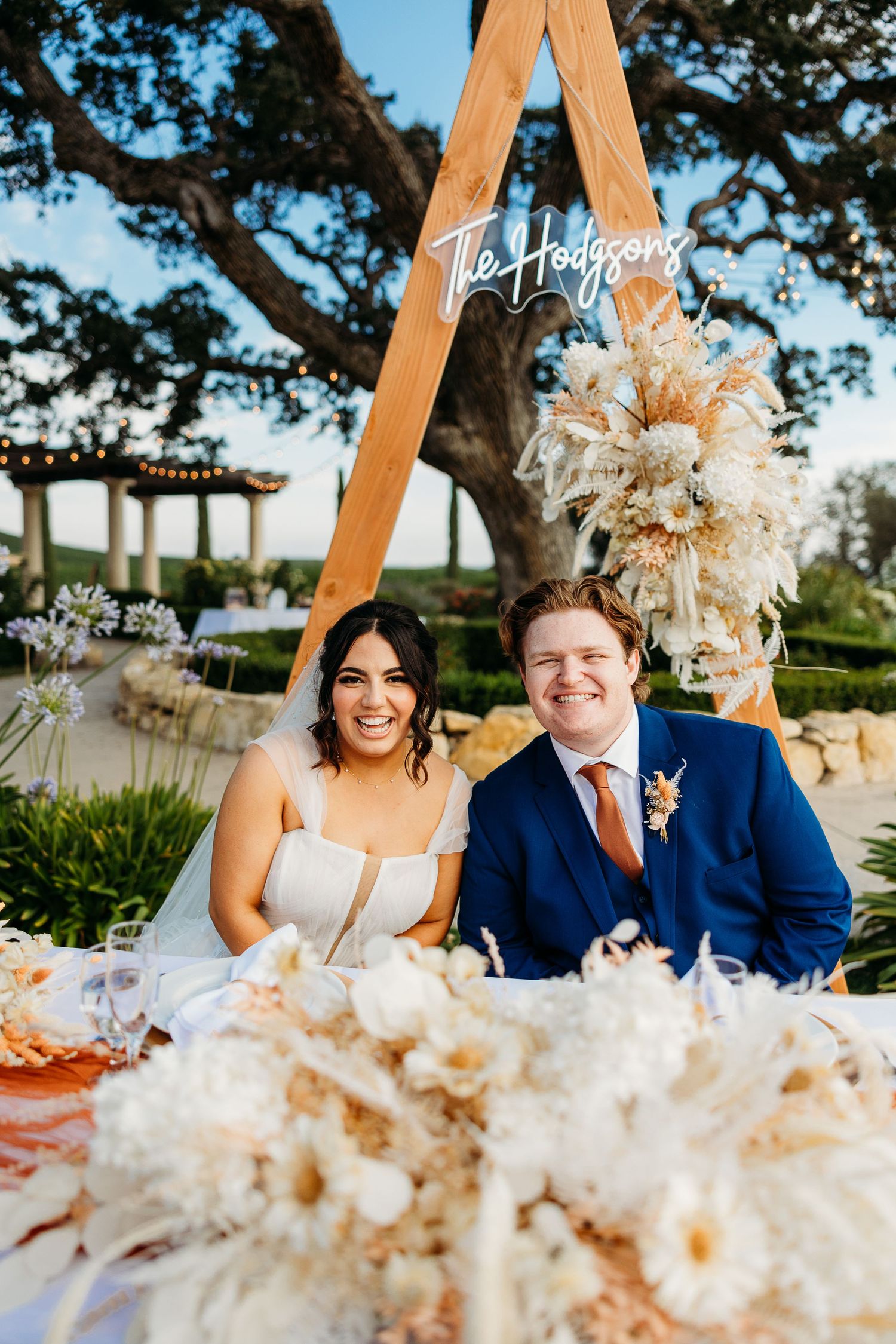 Wedding couple poses at reception table under wooden triangle arch decorated with dried florals.