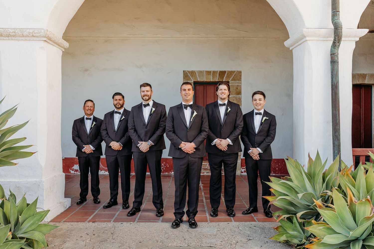 Groomsmen in black tuxedos stand under white arches with agave plants in a Spanish colonial setting.