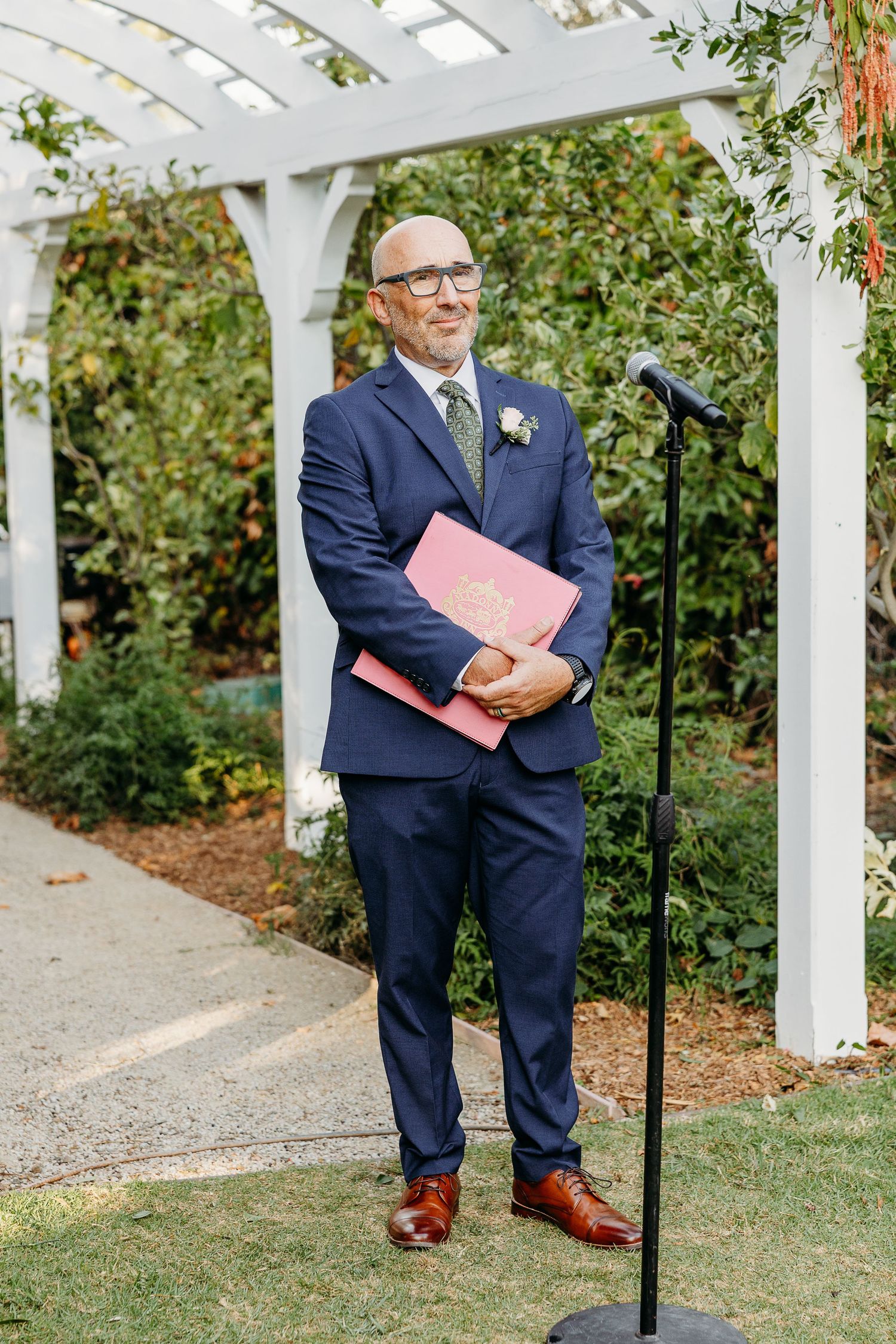 Person in navy suit holds pink folder under white garden pergola.
