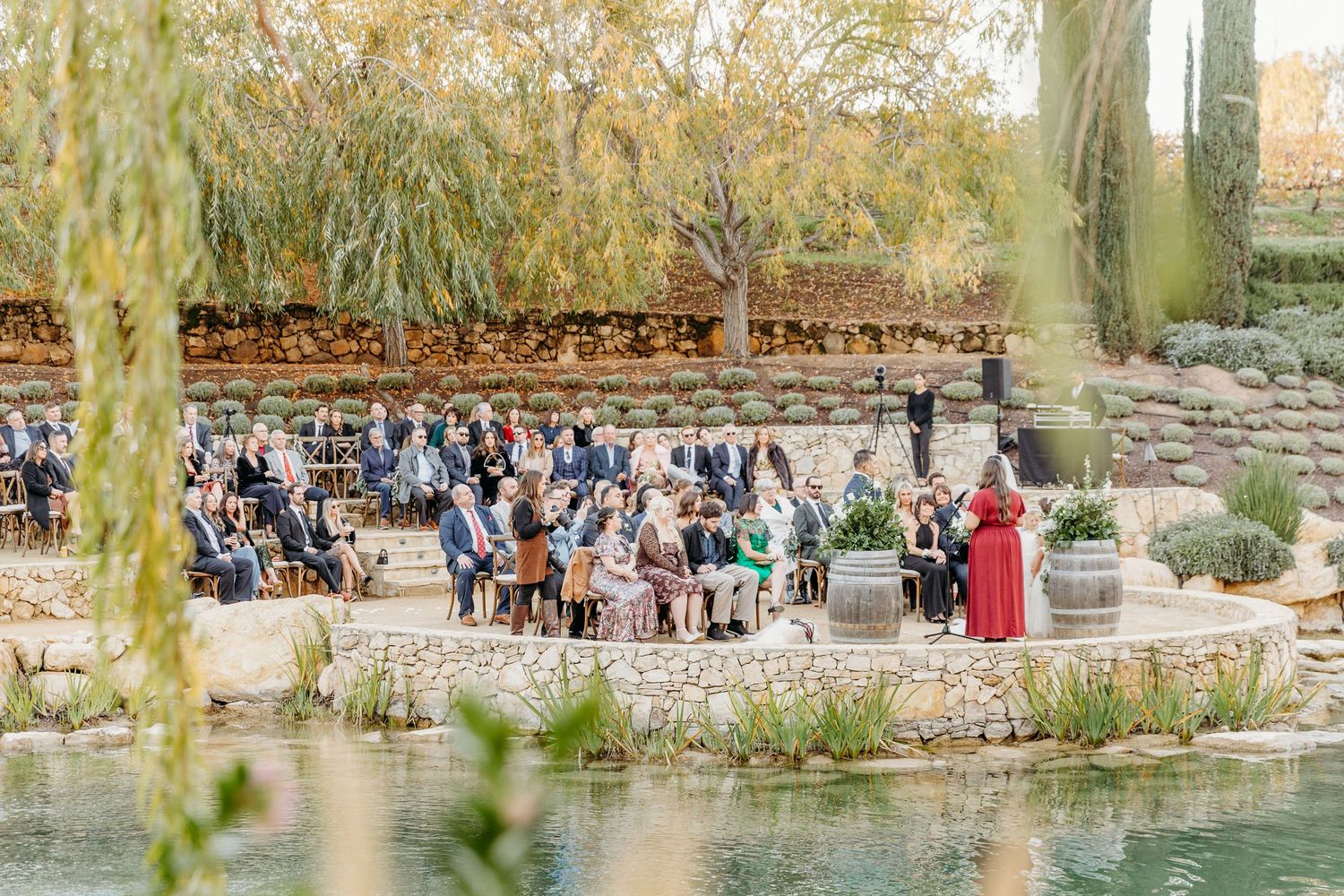 An outdoor wedding ceremony setup with guests seated by a pond under willow trees.