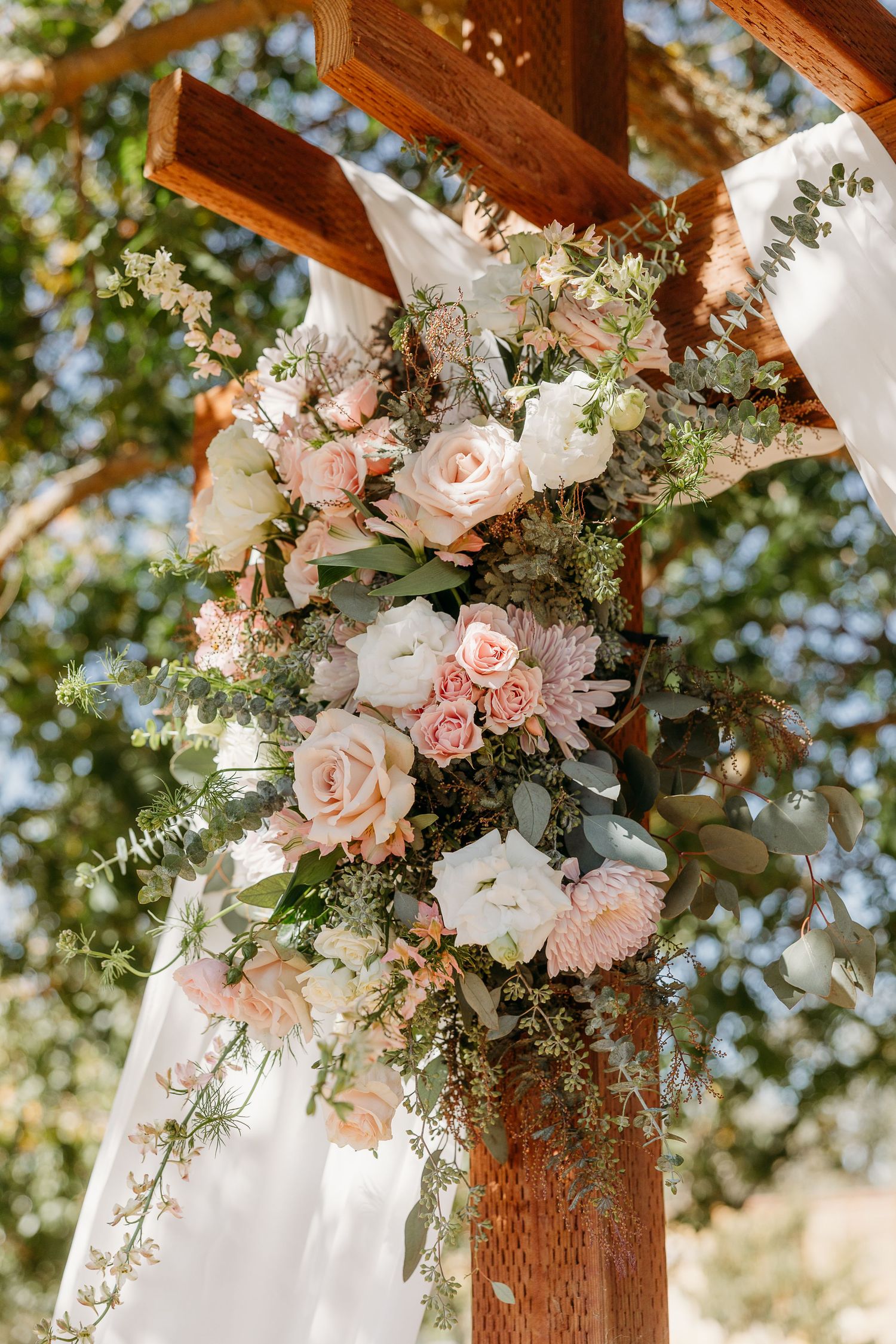 Romantic blush pink and white floral arrangement adorning a wooden wedding arch against a sunny outdoor backdrop.