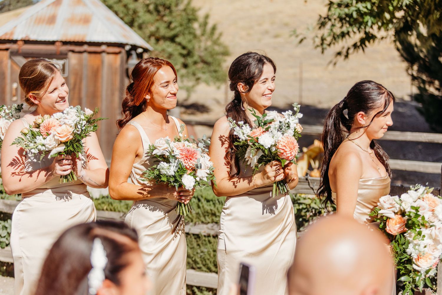 A wedding party in champagne colored dresses stands together holding pink and white floral bouquets in an outdoor setting.