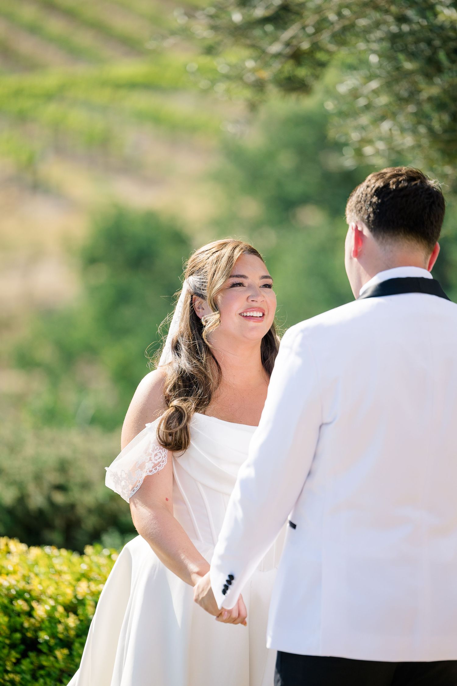 Radiant bride in white off-shoulder gown holds grooms hands smiling during outdoor vineyard wedding ceremony.