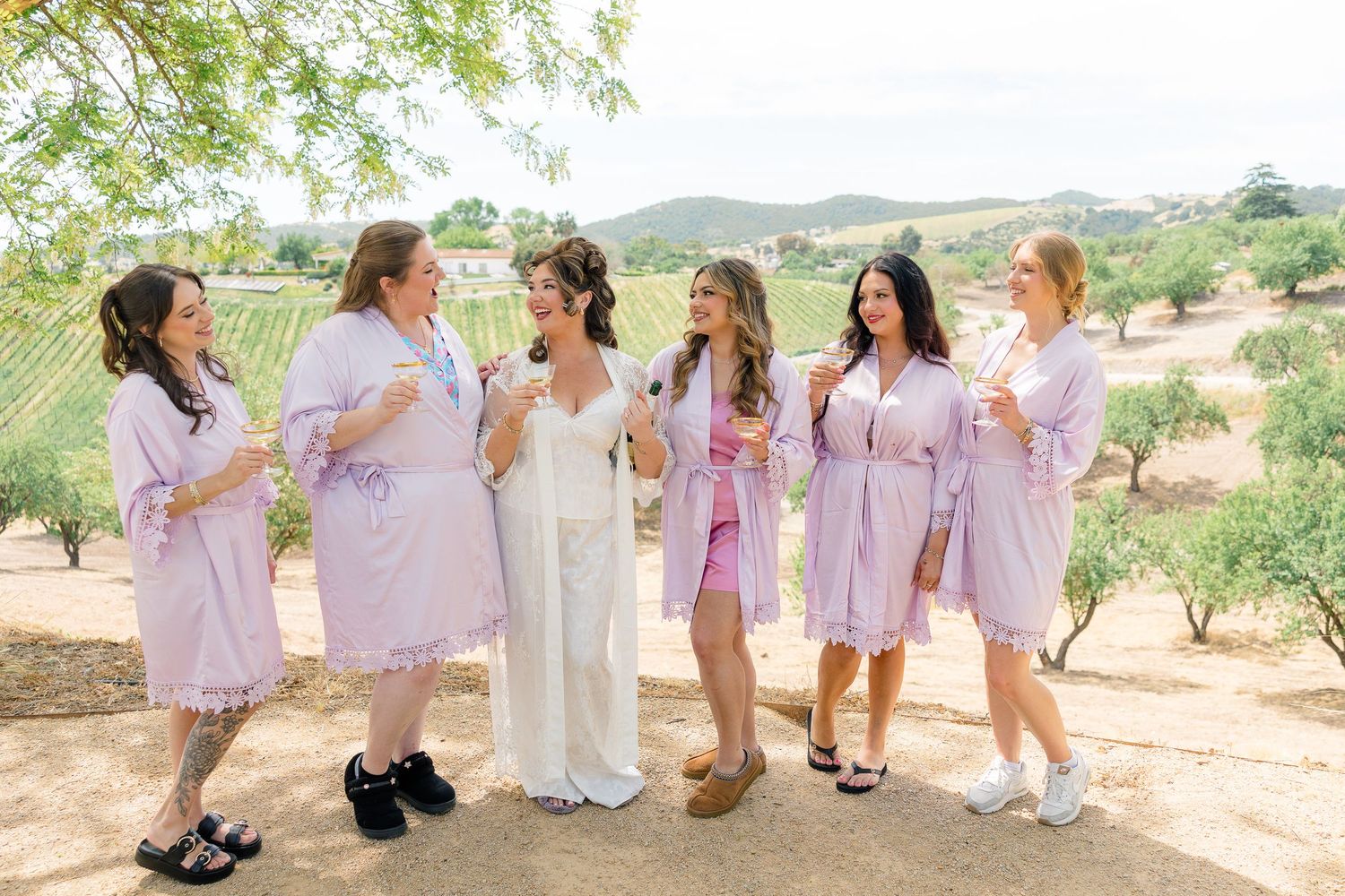 Bride in white gown with five bridesmaids in lavender robes at outdoor vineyard wedding.