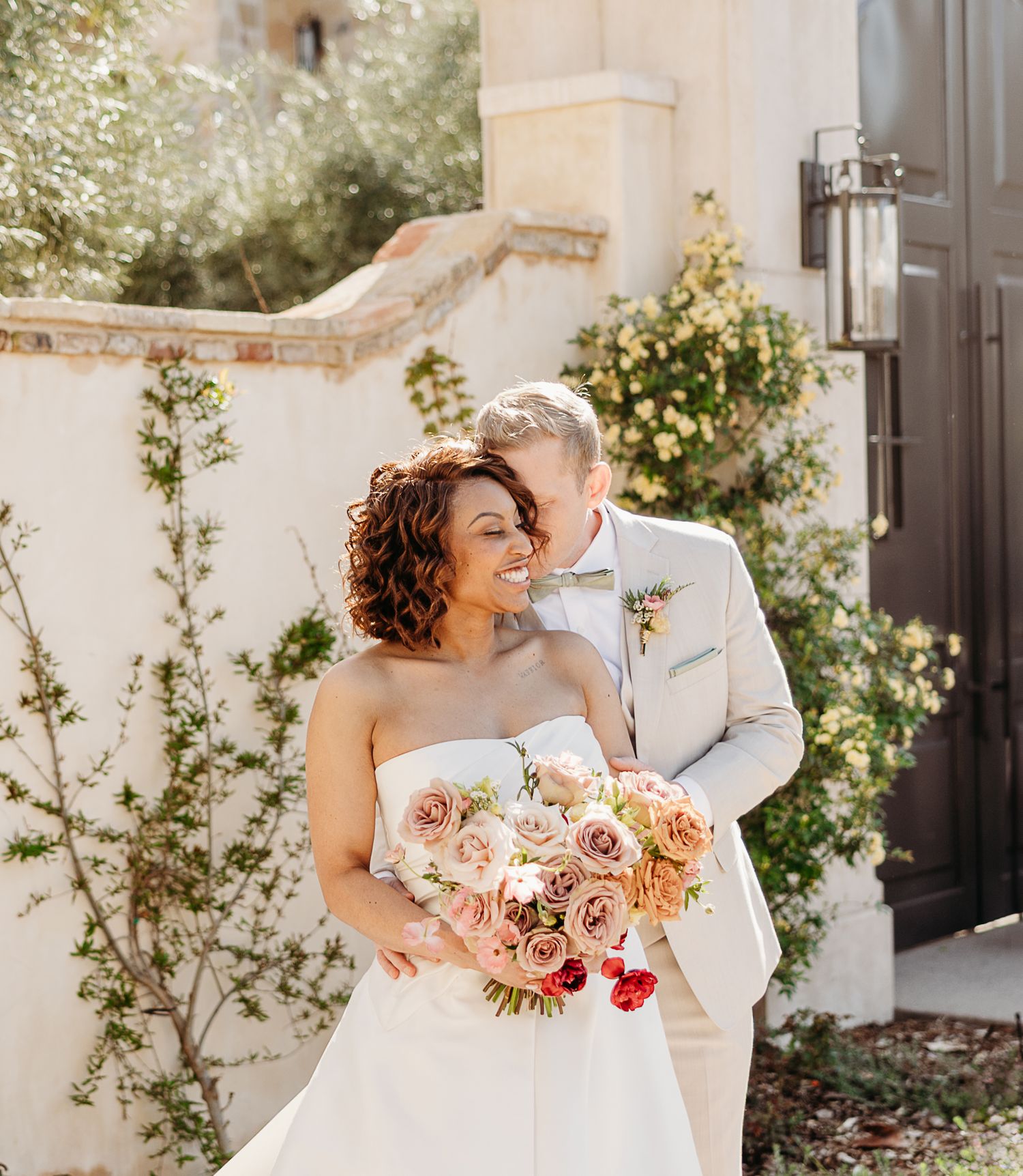Romantic wedding couple shares intimate moments in a sunlit garden with peach-colored floral bouquet.