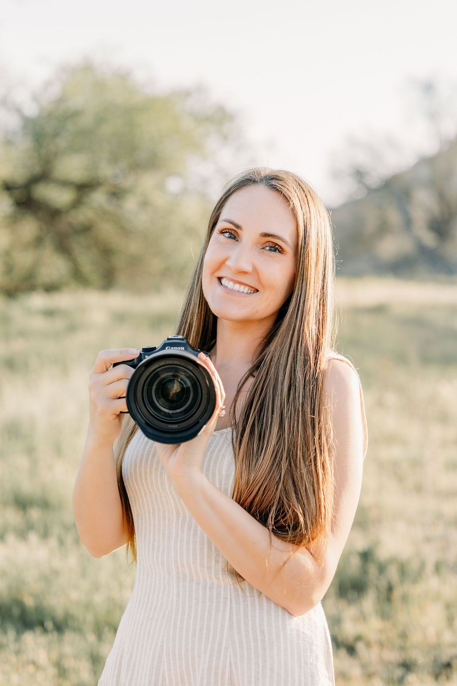 Photographer in white sleeveless dress holds professional camera while posing in sunlit field during golden hour.