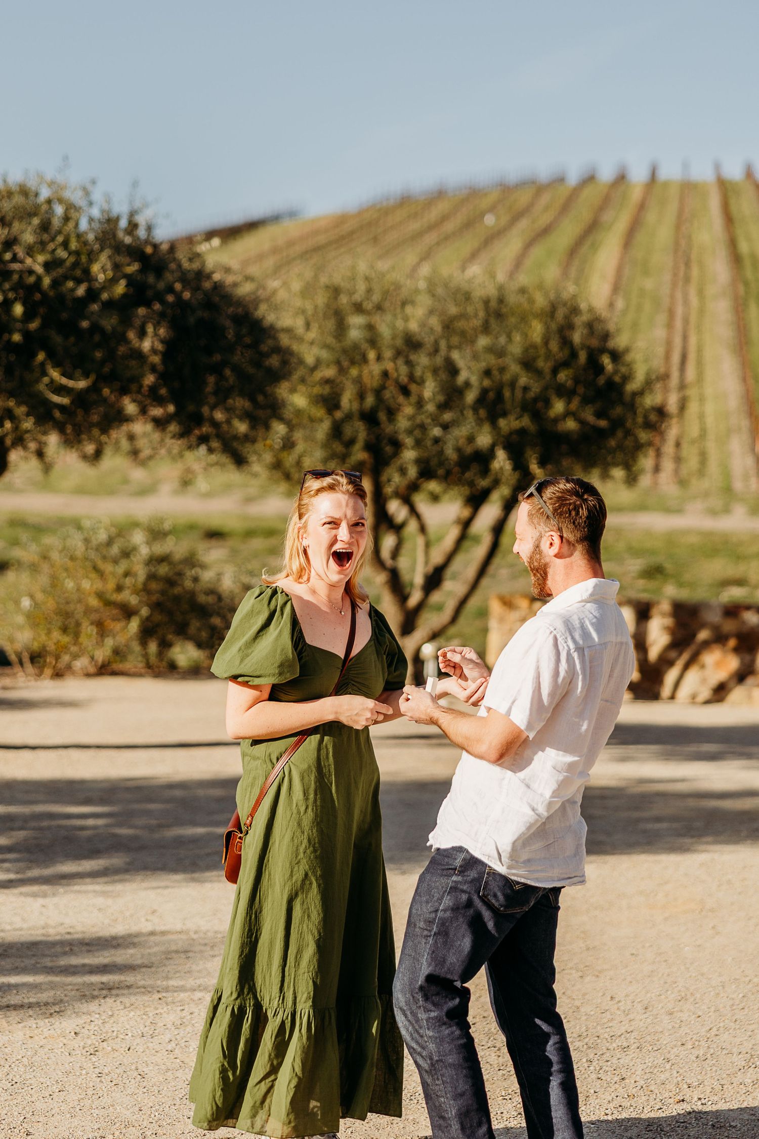 A playful couple in a green dress and white shirt dance together in a vineyard at sunset.