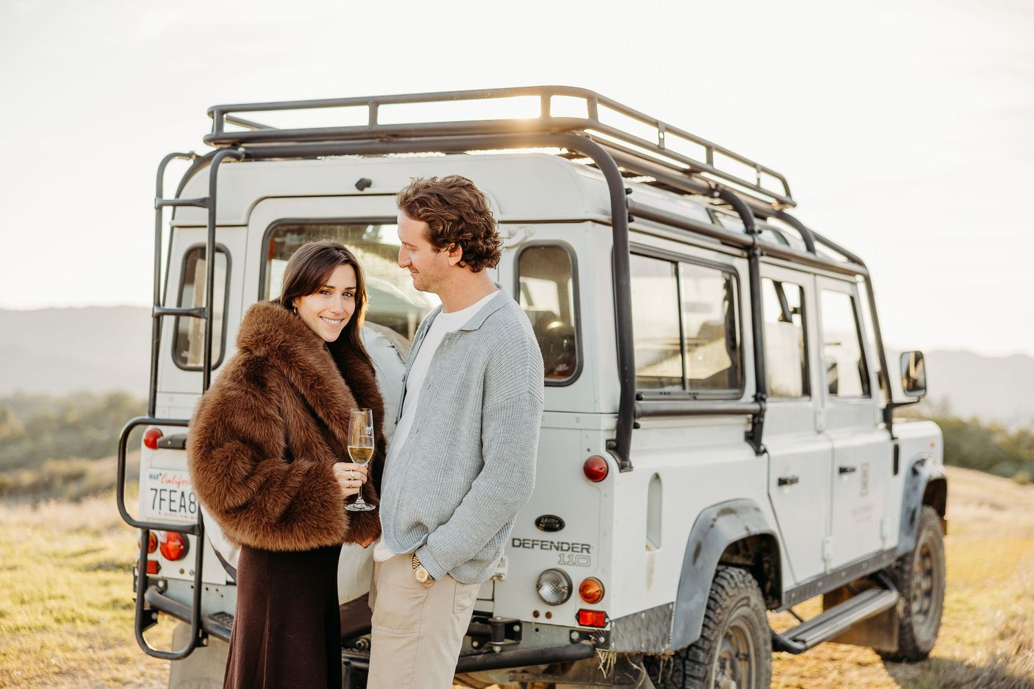 Romantic couple sharing tender moments near a vintage Land Rover Defender during a golden hour photoshoot in nature.