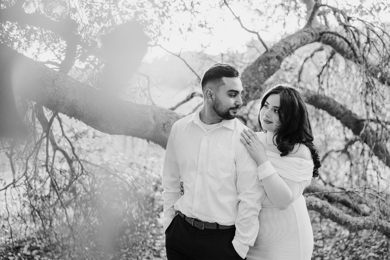 A couple shares a tender moment together under a tree branch in black and white photography.