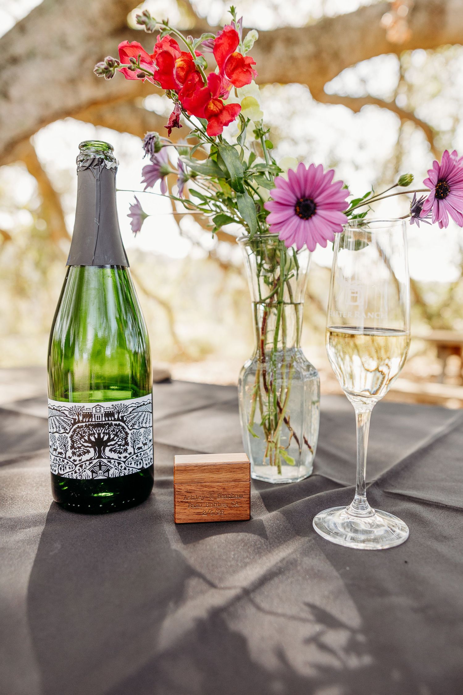 A wine bottle and glass sit beside a vase of red and purple flowers on a table with dappled sunlight.
