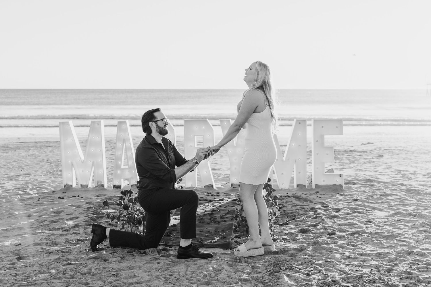 A romantic beach proposal scene with white letters spelling out 'MARRY ME' arranged in the sand at sunset.