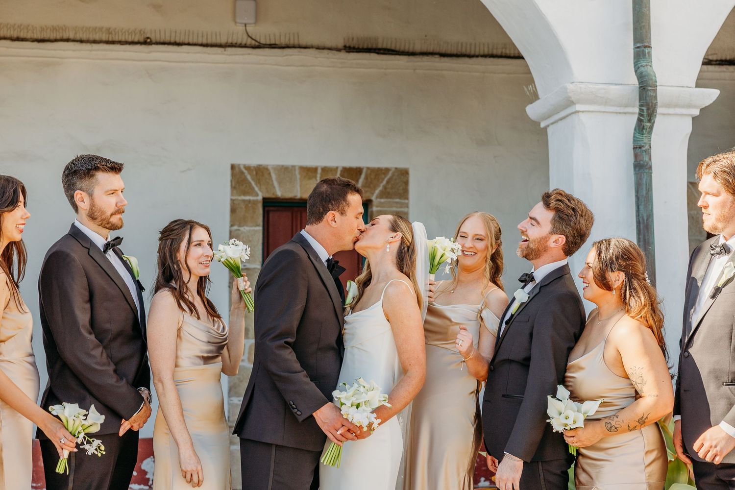 Wedding party poses for candid photo series under white arched walkway.