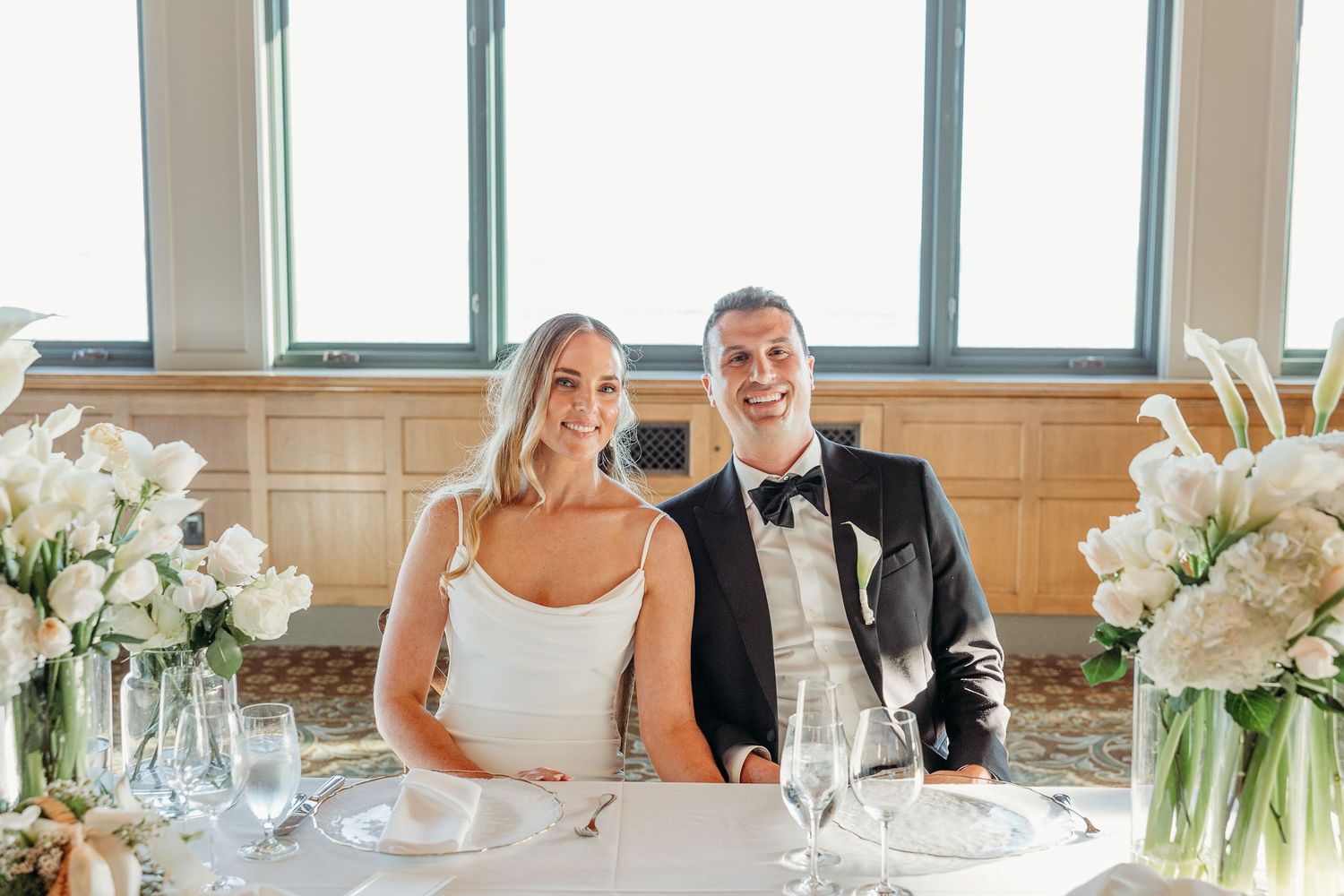 A couple sits at their wedding reception table decorated with white floral arrangements.