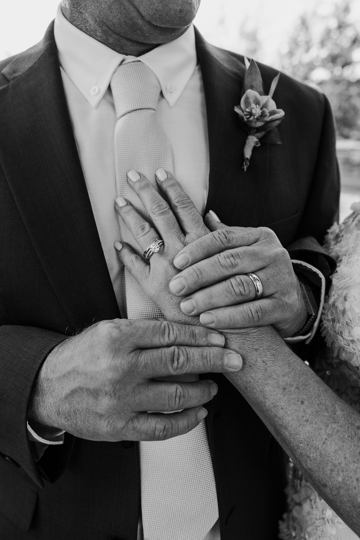A black and white close-up of hands with wedding rings and a boutonniere on a suit lapel.
