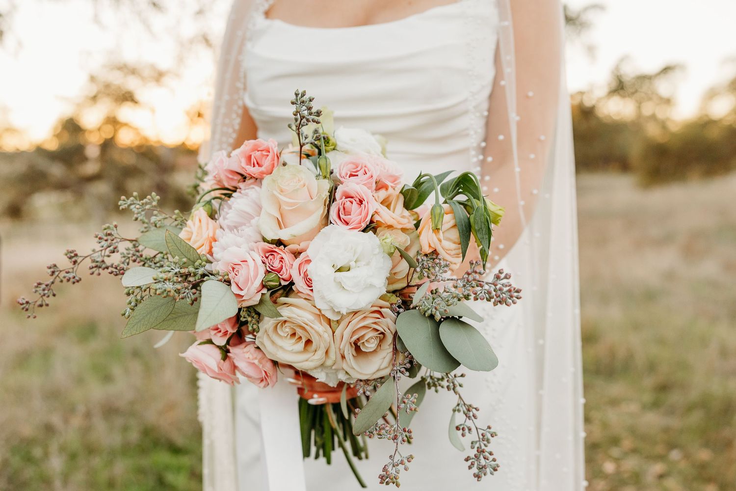 A bridal bouquet featuring blush pink roses and eucalyptus leaves against a sunlit outdoor backdrop.