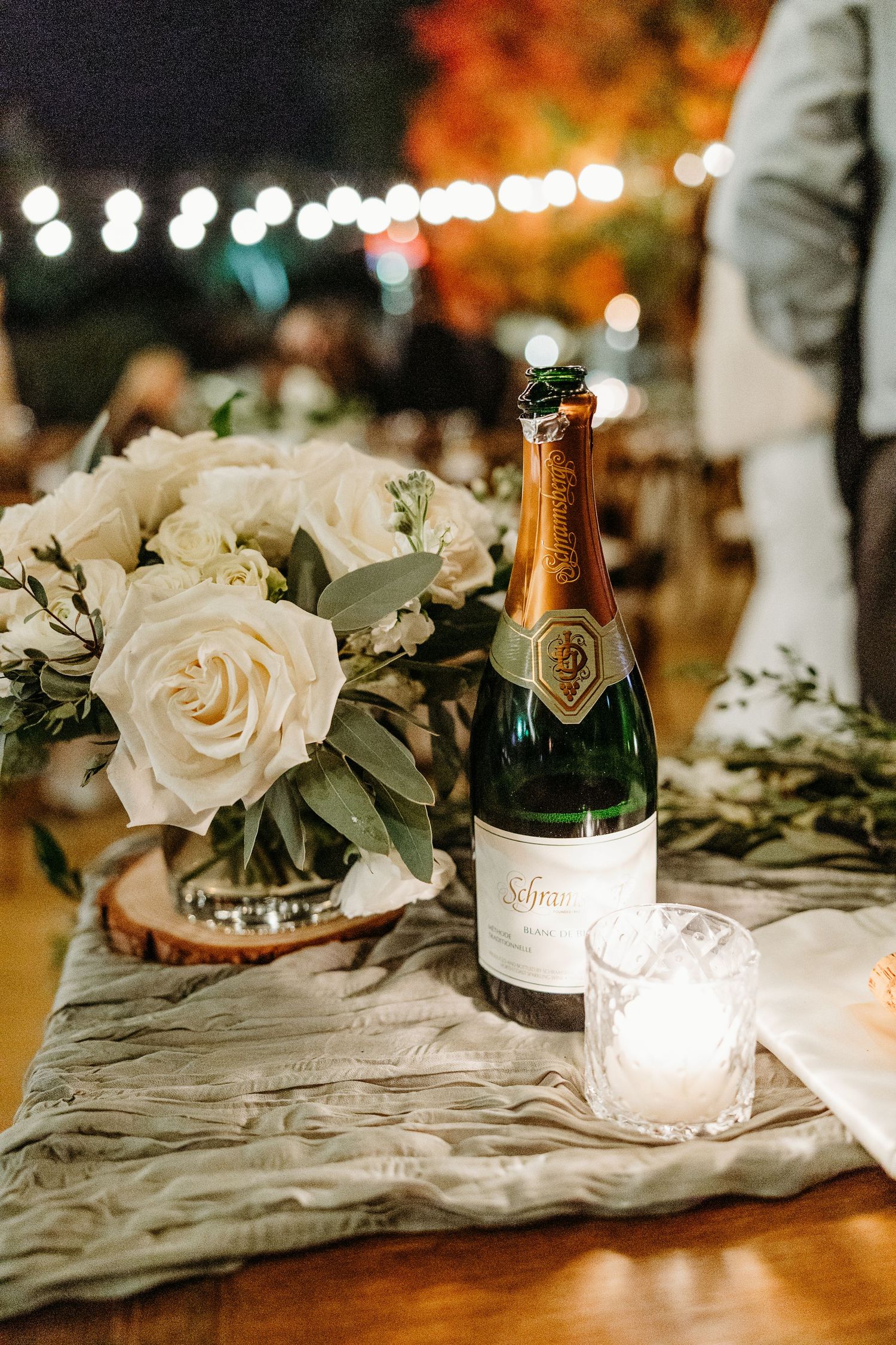 Elegant wedding table decor with white roses, champagne bottles and string lights creating a romantic ambiance.