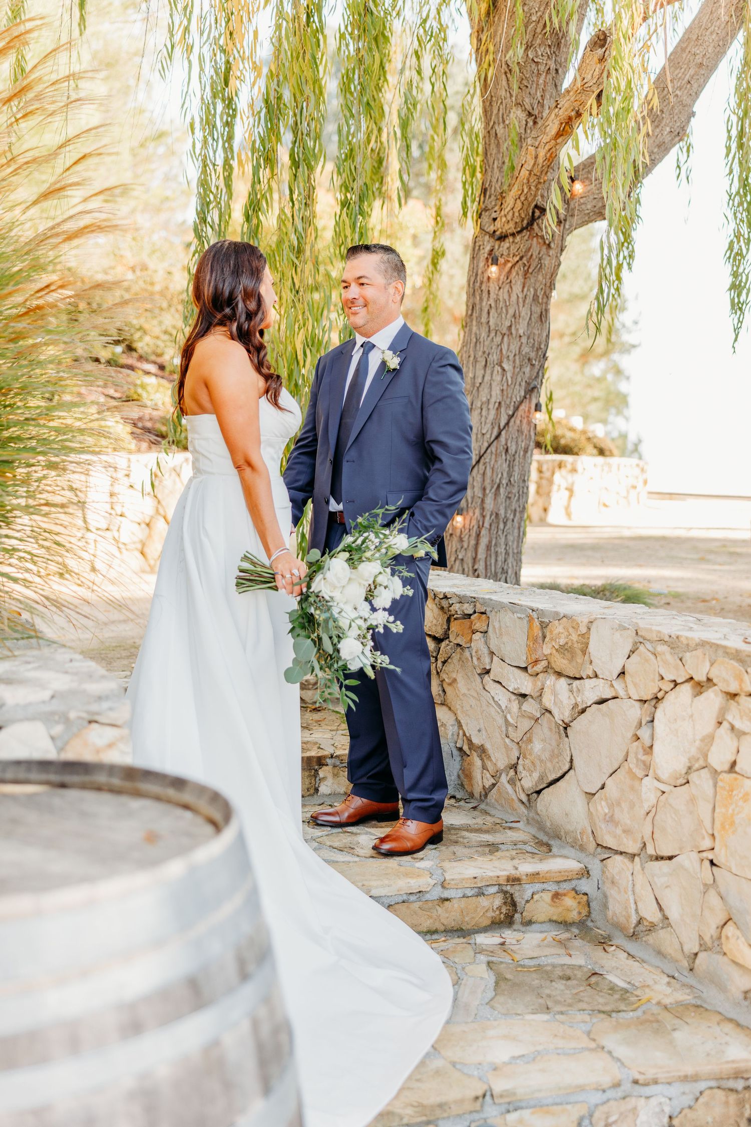 Elegant wedding couple shares an intimate moment under a willow tree by a stone wall.