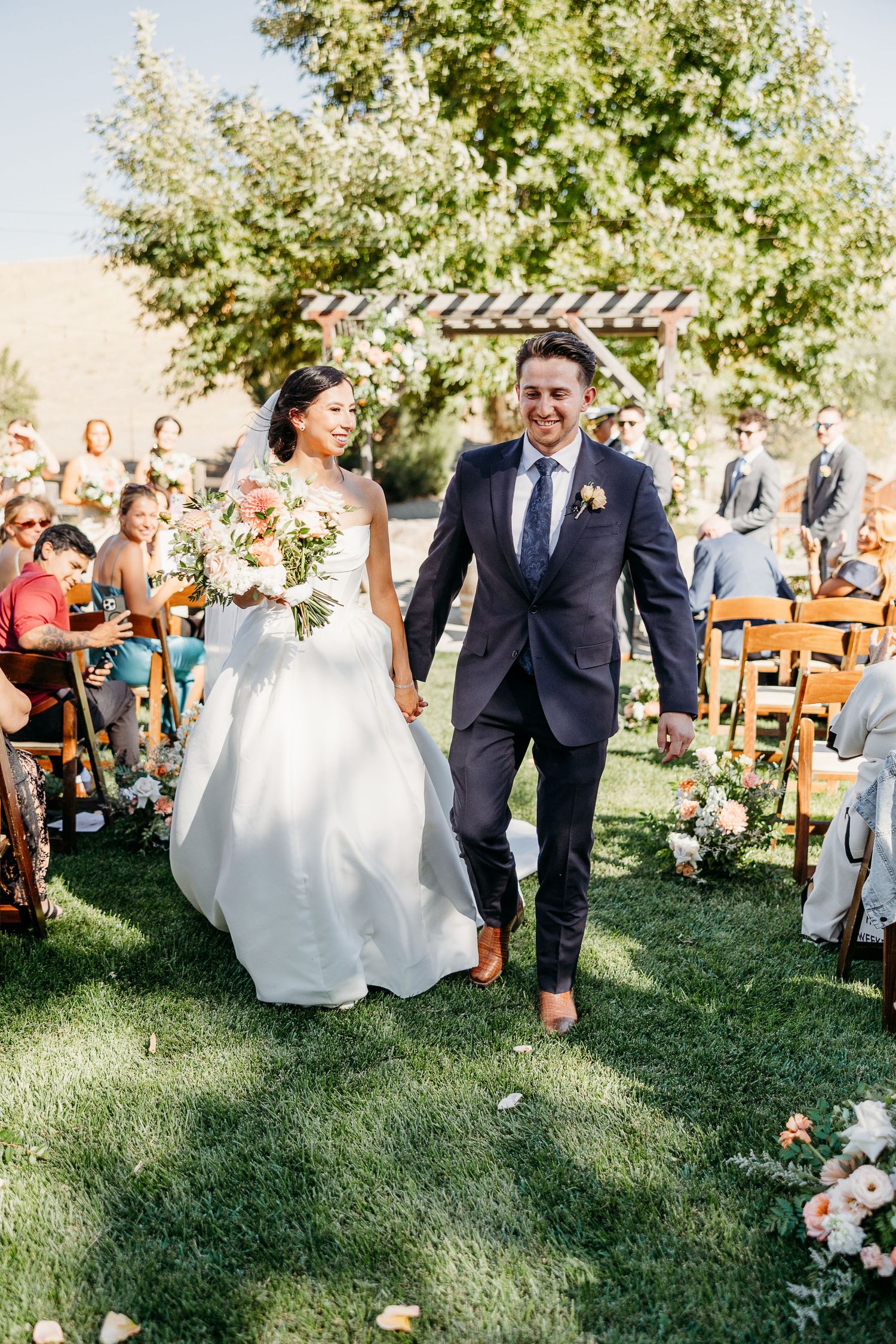 Wedding party walks down grassy aisle as guests toss flower petals during outdoor summer ceremony celebration.