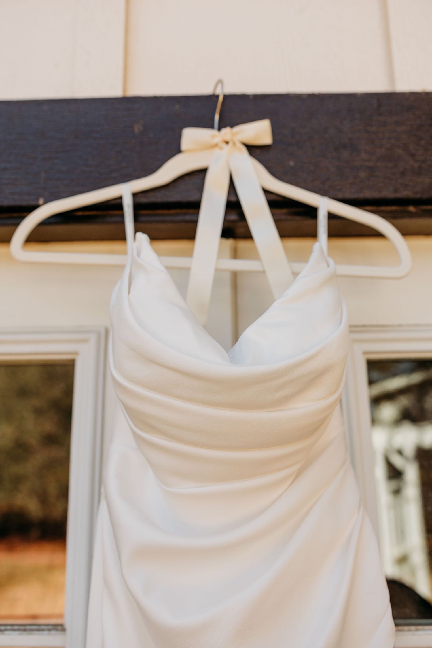 A white wedding dress with ruched bodice hangs on a white plastic hanger with a decorative bow detail.