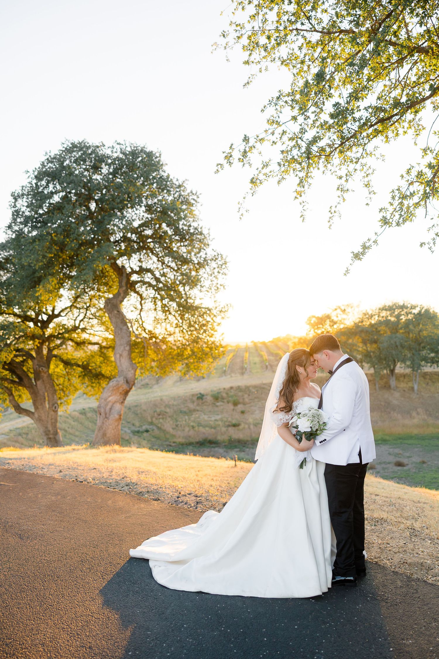Bride and groom share romantic sunset kiss on tree-lined road, wedding dress trailing.