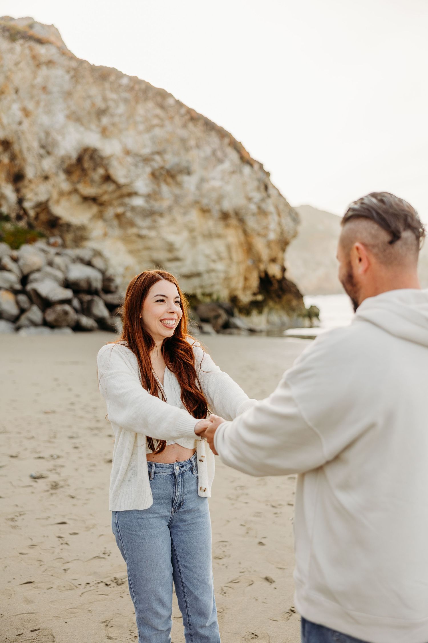 Two people holding hands and sharing a joyful moment on a beach with dramatic coastal cliffs in the background.