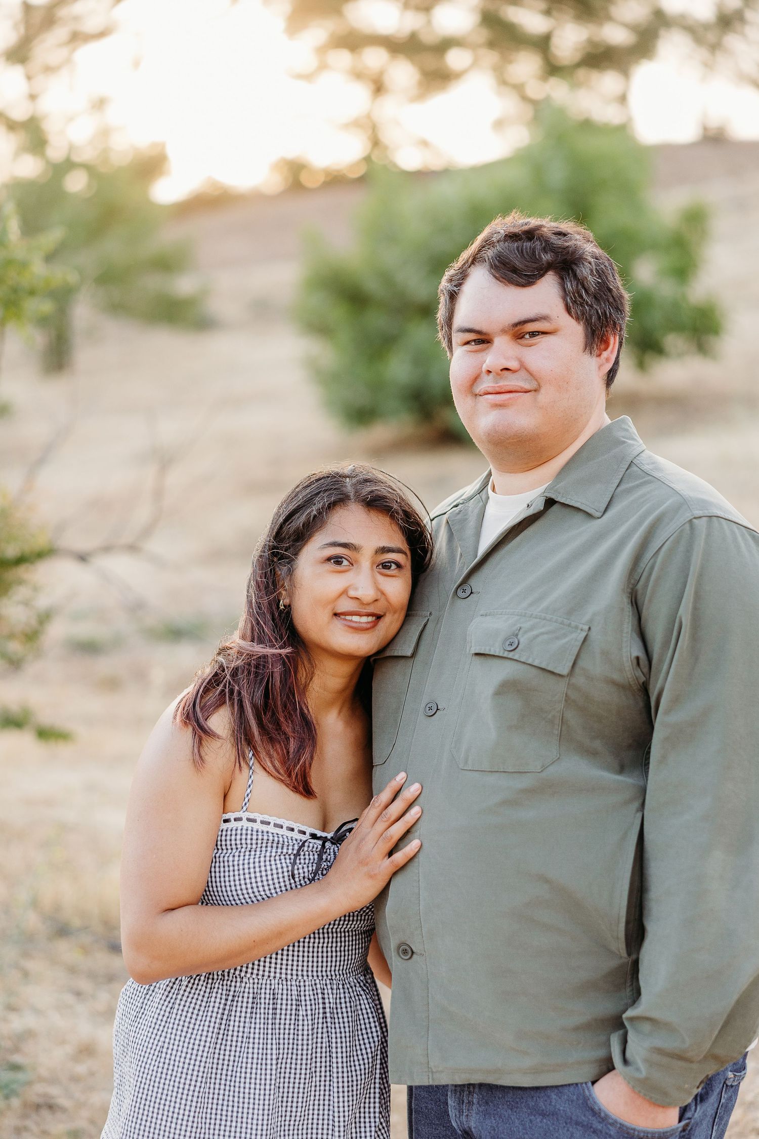 A romantic couple embraces in a desert setting during golden hour, wearing casual attire in gingham and olive tones.