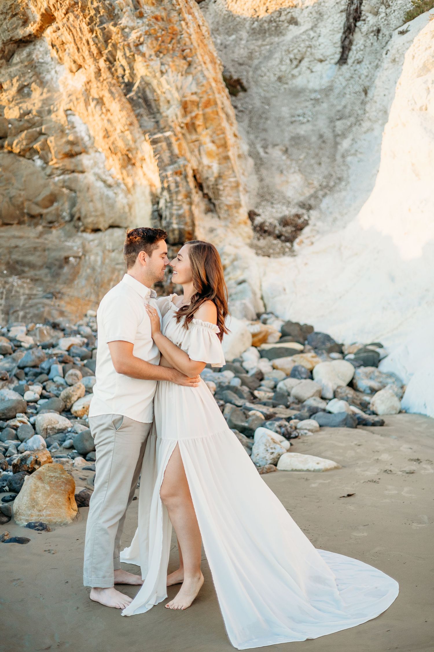 Couple in elegant white outfits dancing on beach with rocky cliffs and waves behind them.