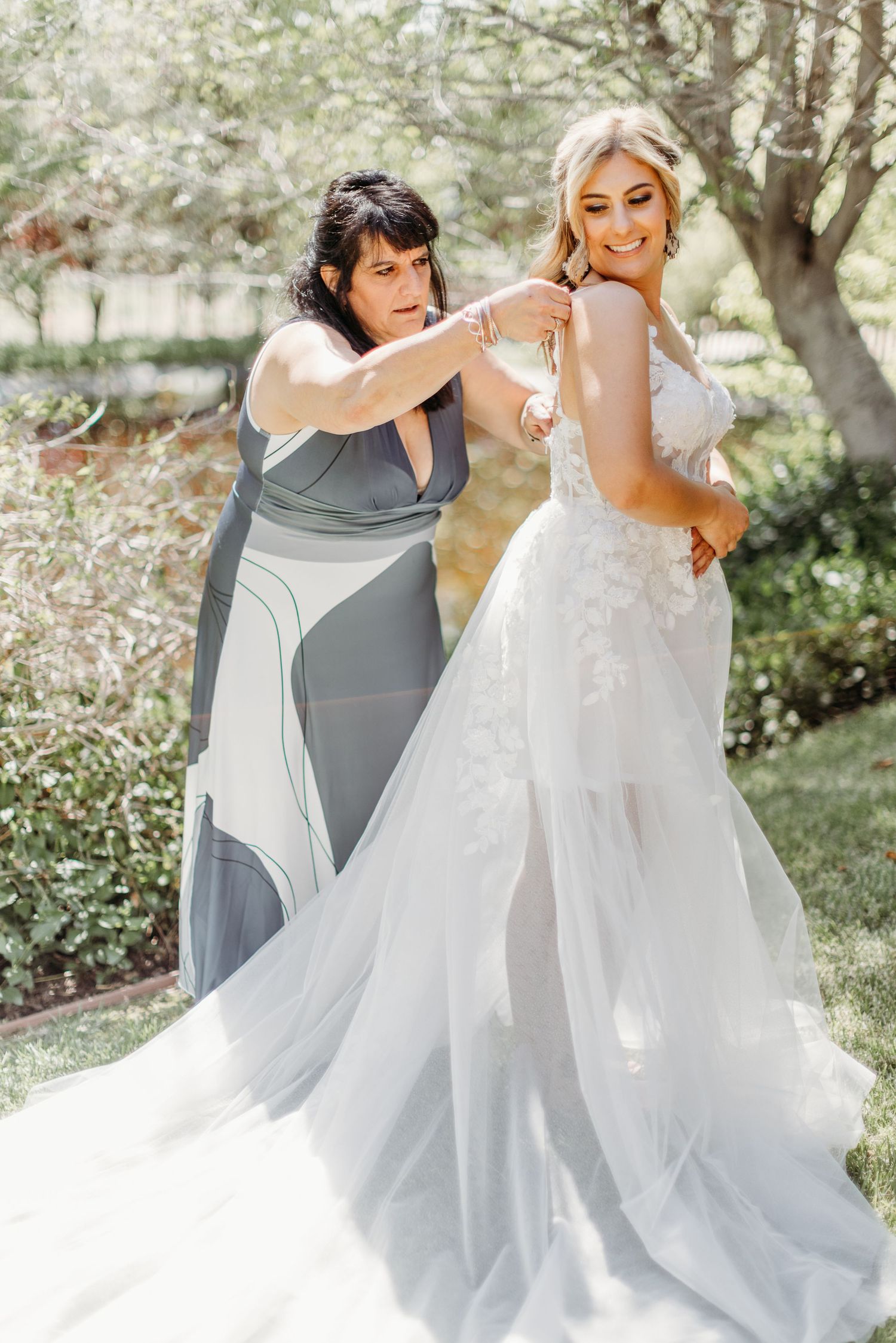 A bride in a white lace gown gets final adjustments made to her dress outdoors in a garden setting.