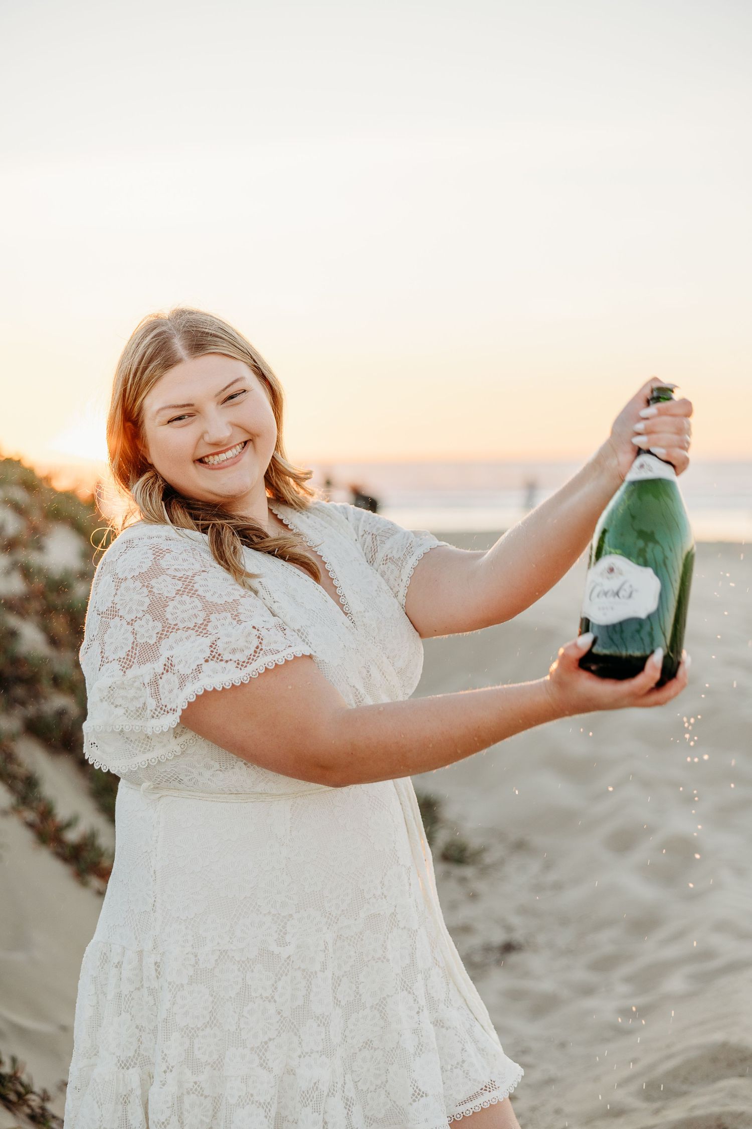 Graduate in white dress celebrates by popping champagne on beach during golden hour.
