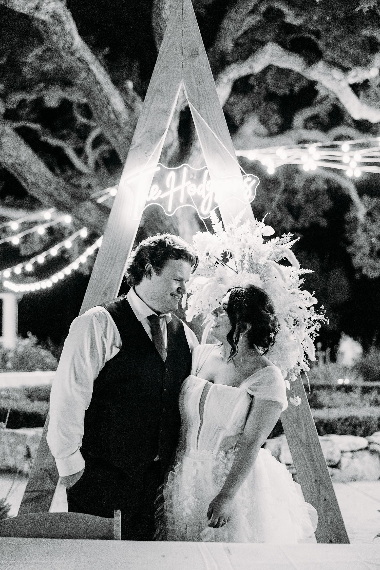 Black and white photo of couple beneath illuminated wedding arch decorated with dried flowers.