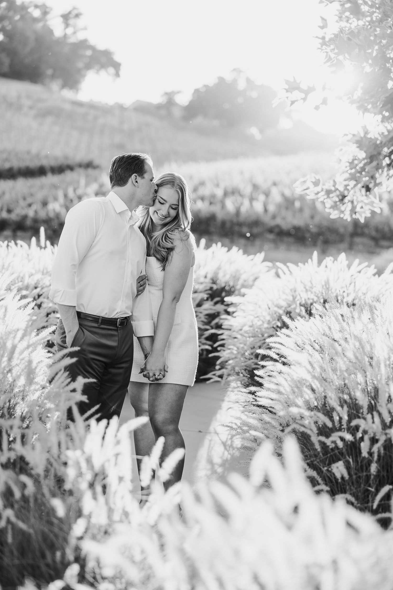 Black and white photo of couple walking through tall pampas grass during outdoor photoshoot.