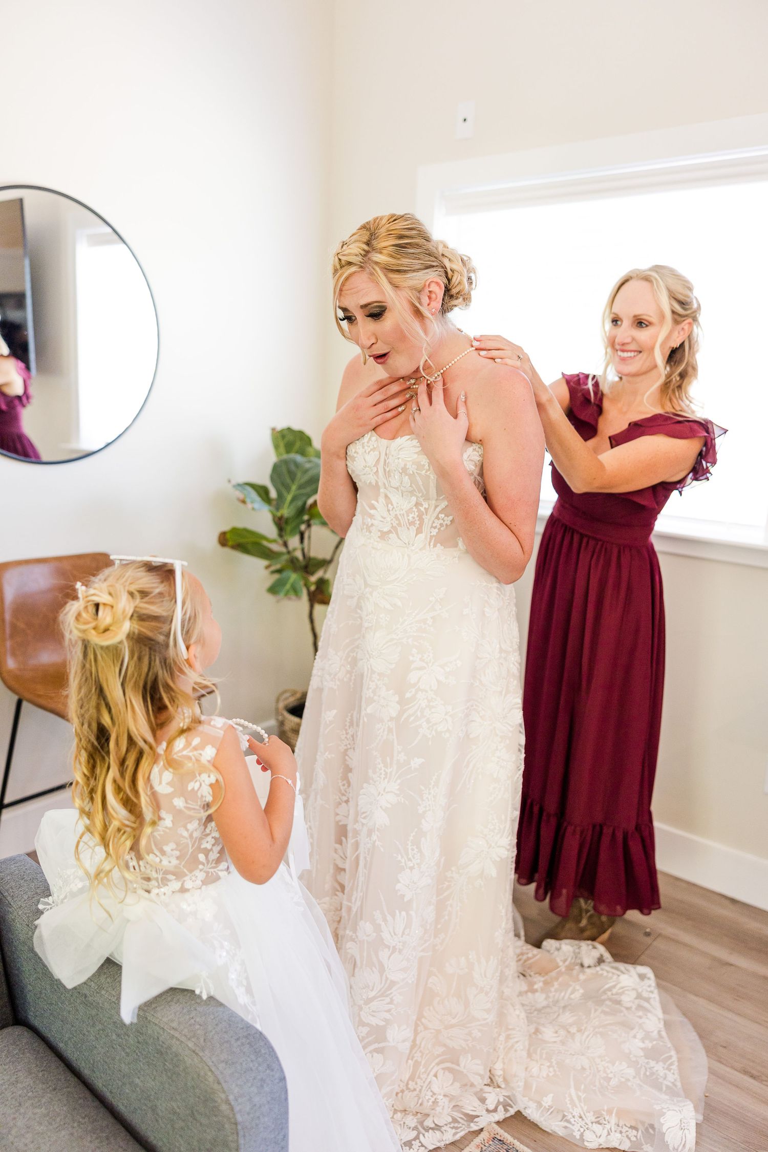 A touching pre-wedding moment as bride in lace dress interacts with wedding party in sunlit room.