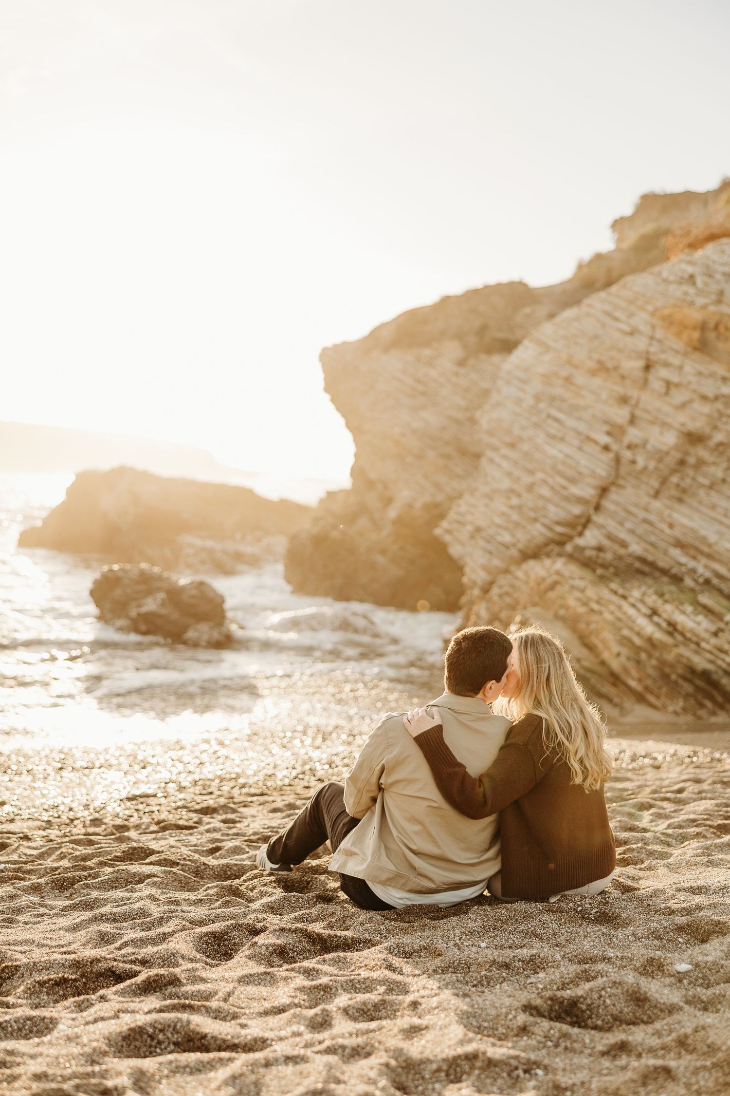 A couple relaxes together on a sandy beach at sunset with rocky coastal cliffs in the background.