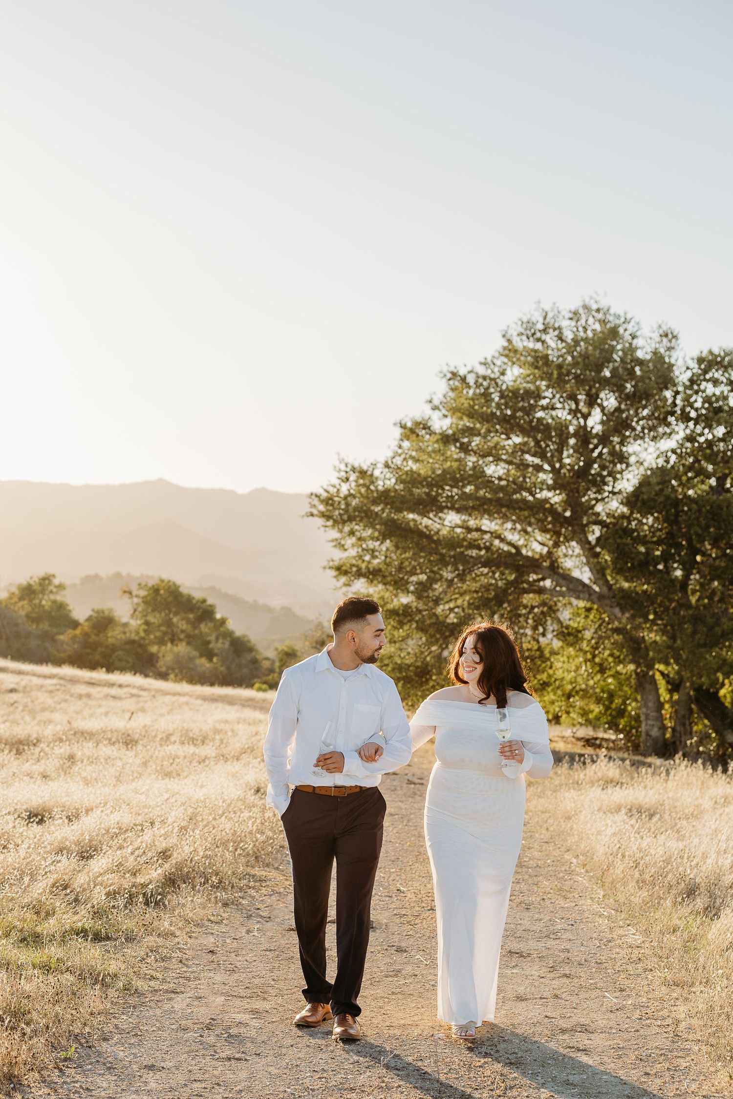 A couple in white outfits walks together through a golden field at sunset with mountains in the background.