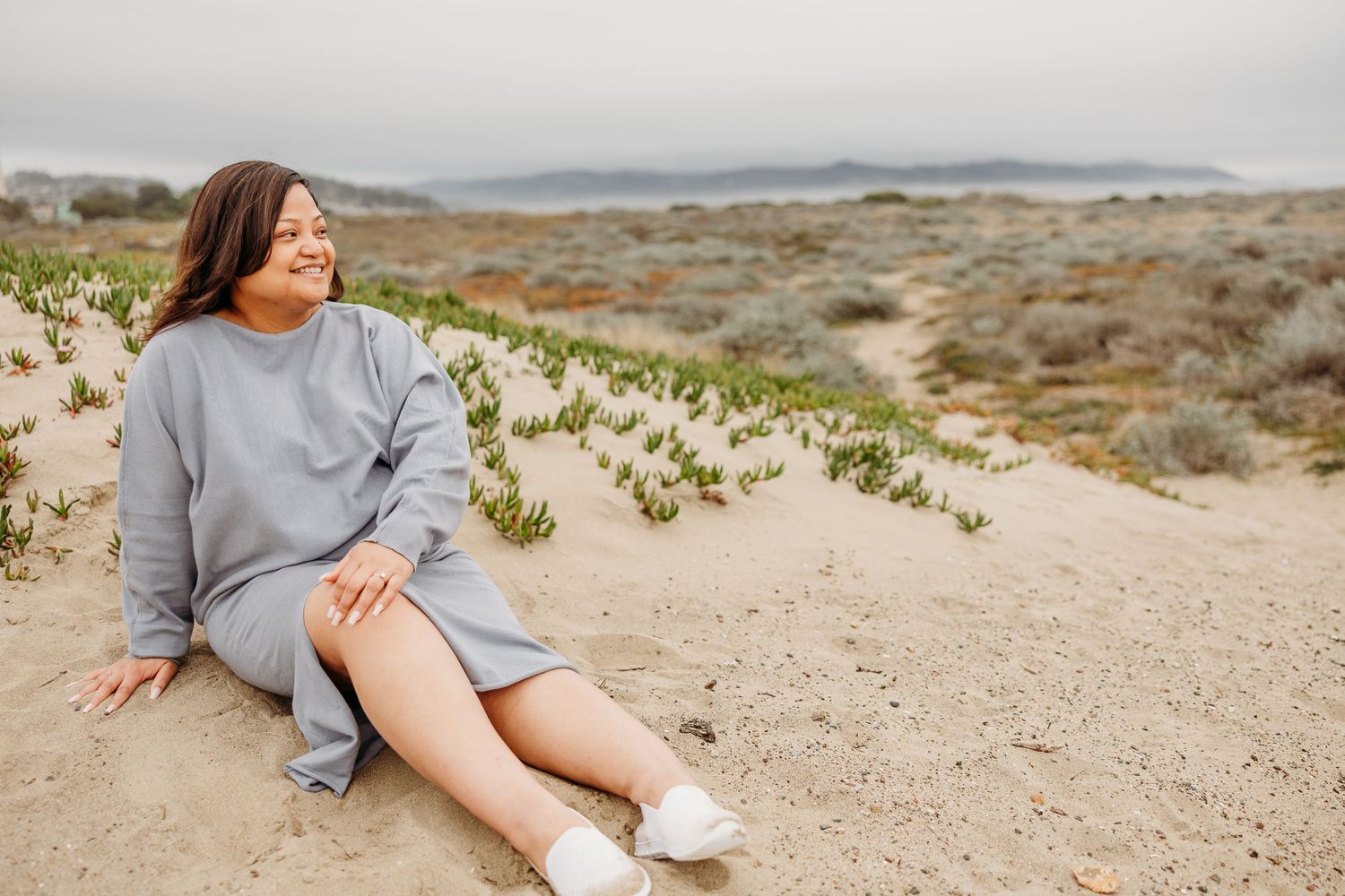 Person in gray sweater dress relaxing on sandy dunes with coastal vegetation and mountains in background.