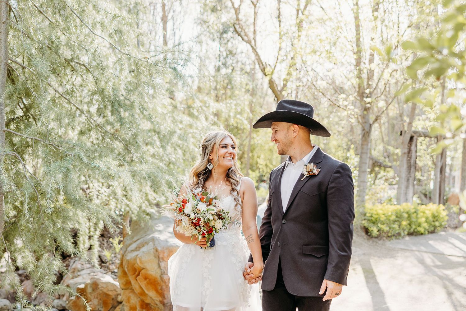 A bride and groom in western-style wedding attire walking hand in hand along a garden path.