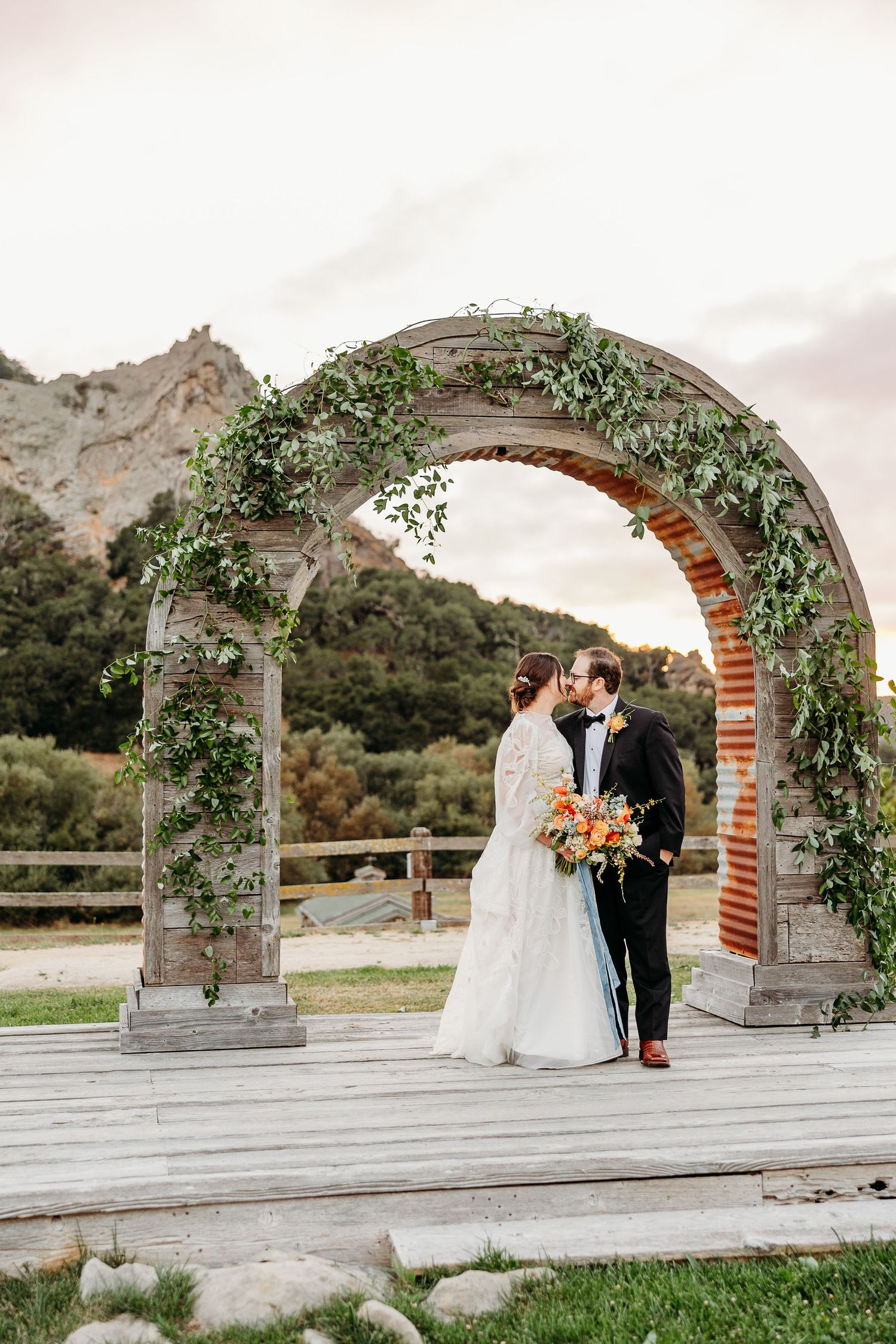 A romantic wedding couple shares a moment under an elegant stone archway decorated with greenery and florals best SLO wedding photographer.