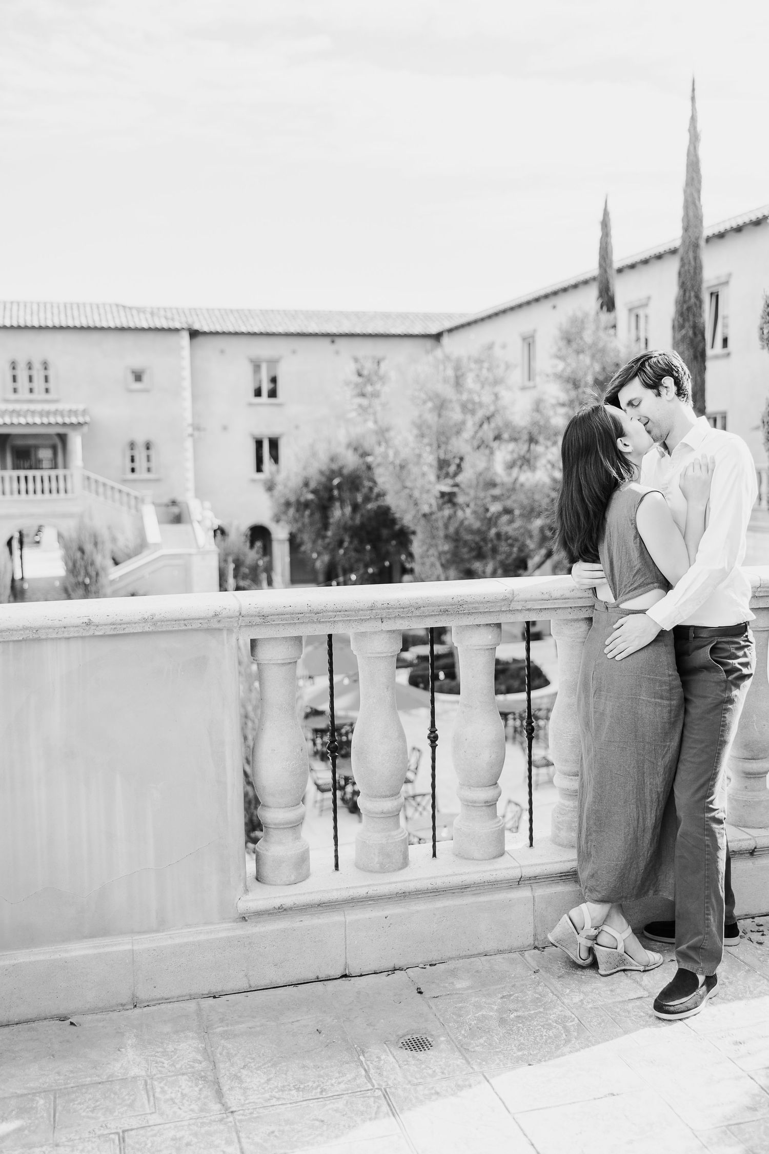 A romantic black and white photograph of a couple embracing on a balcony with Mediterranean architecture in the background