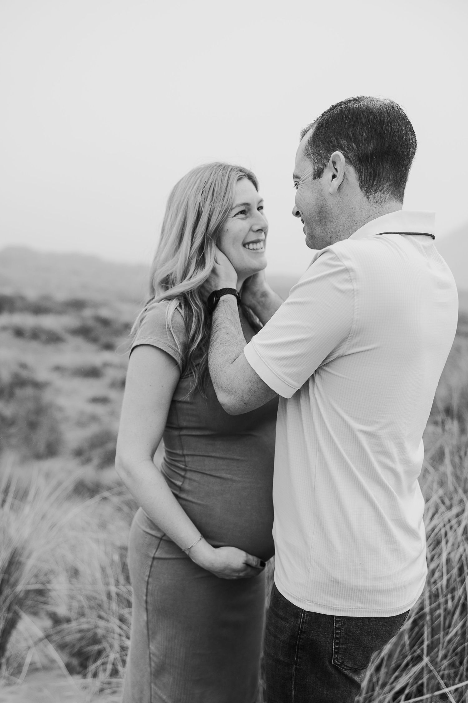 A tender black and white portrait of an expectant couple sharing an intimate moment among coastal dunes.