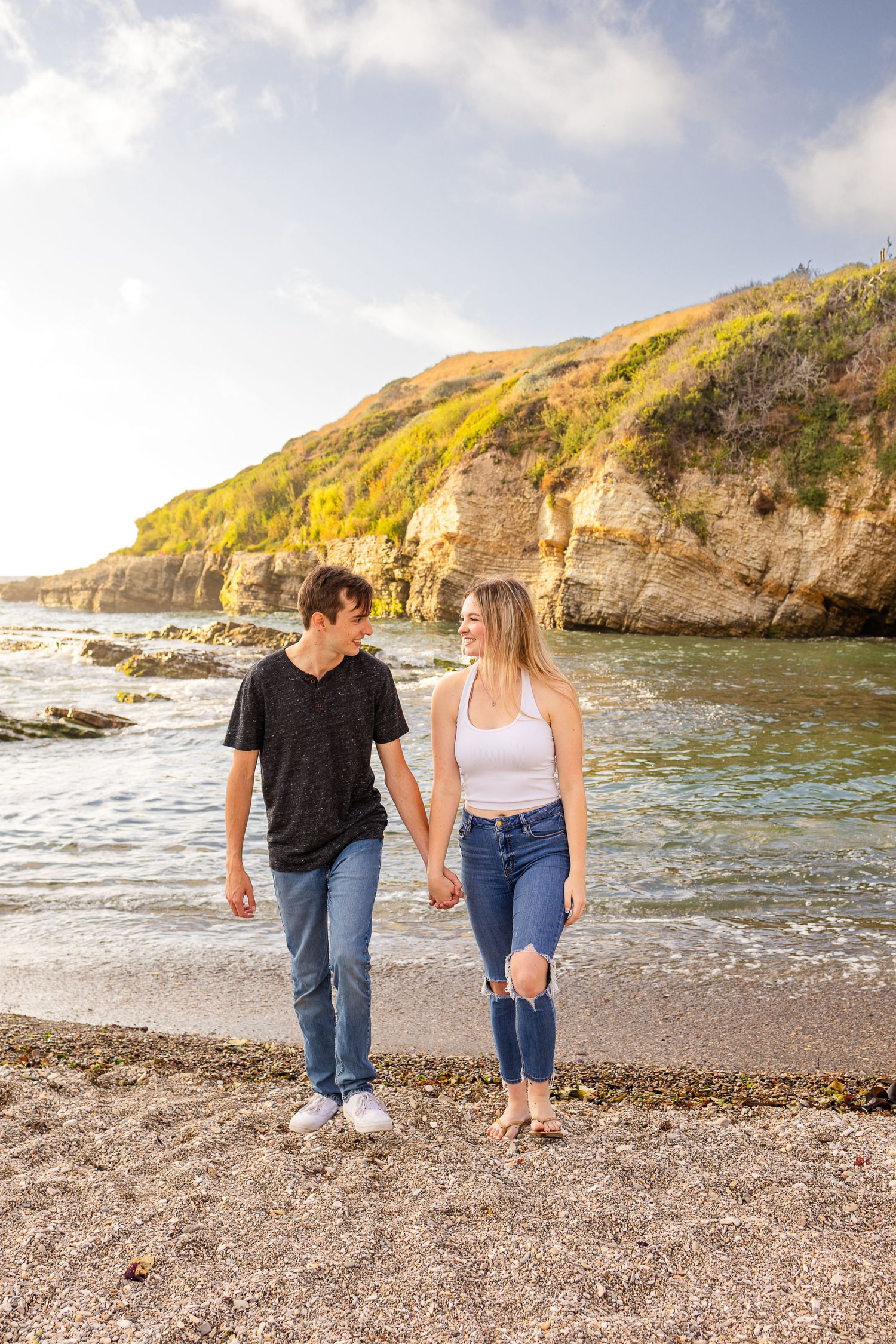 A couple strolls together on a pebble beach with dramatic coastal cliffs behind them.