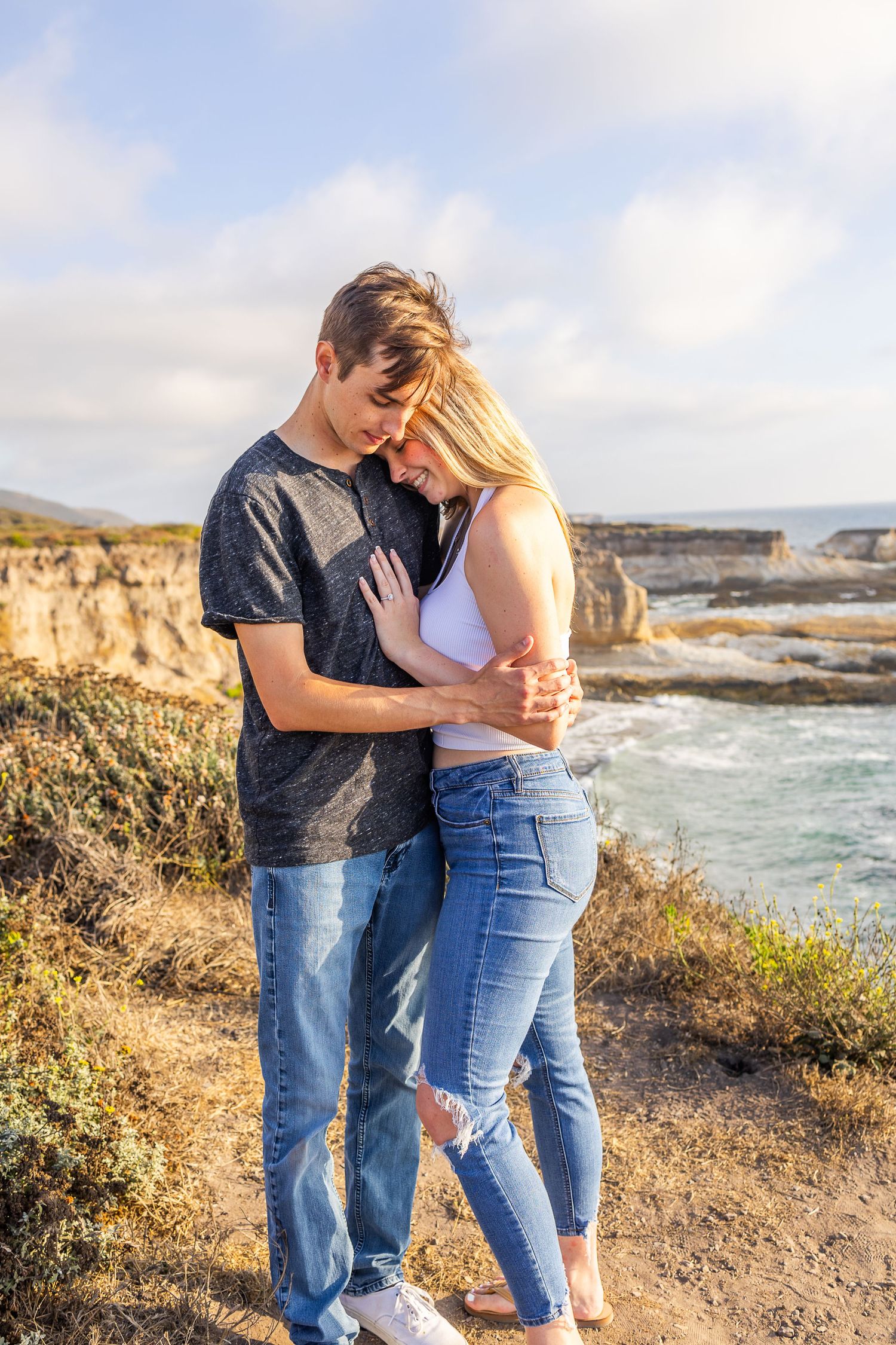 A romantic couple embraces on a cliff edge with crashing waves and rocky coastline in the background.