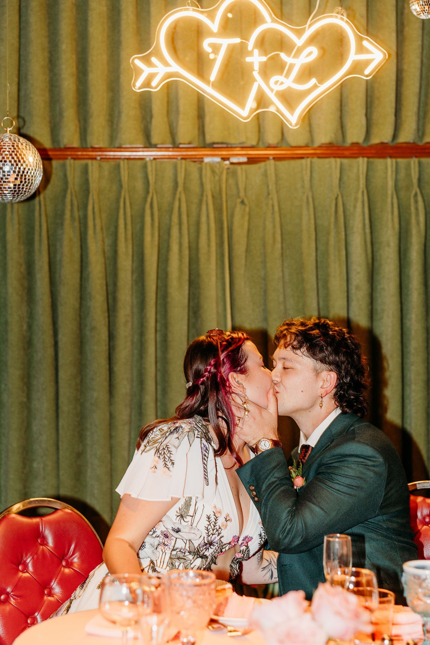 A romantic moment under a neon heart sign with green curtain backdrop at a wedding reception.