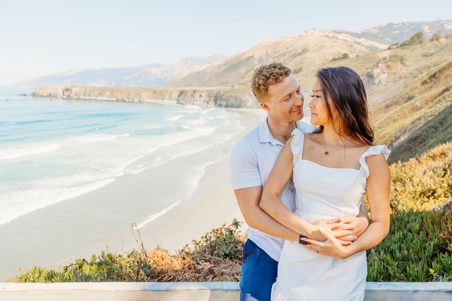 A romantic embrace overlooking a dramatic California coastline with waves and cliffs.