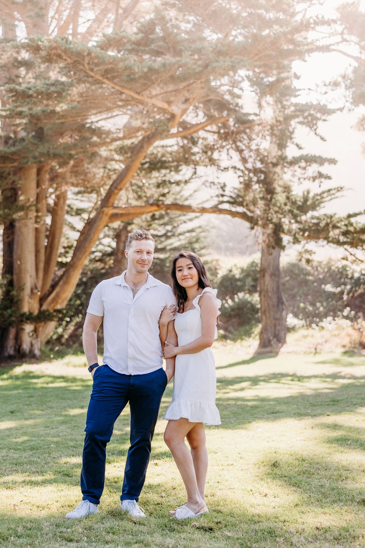 Happy couple poses together under tall pine trees in sunny park setting.