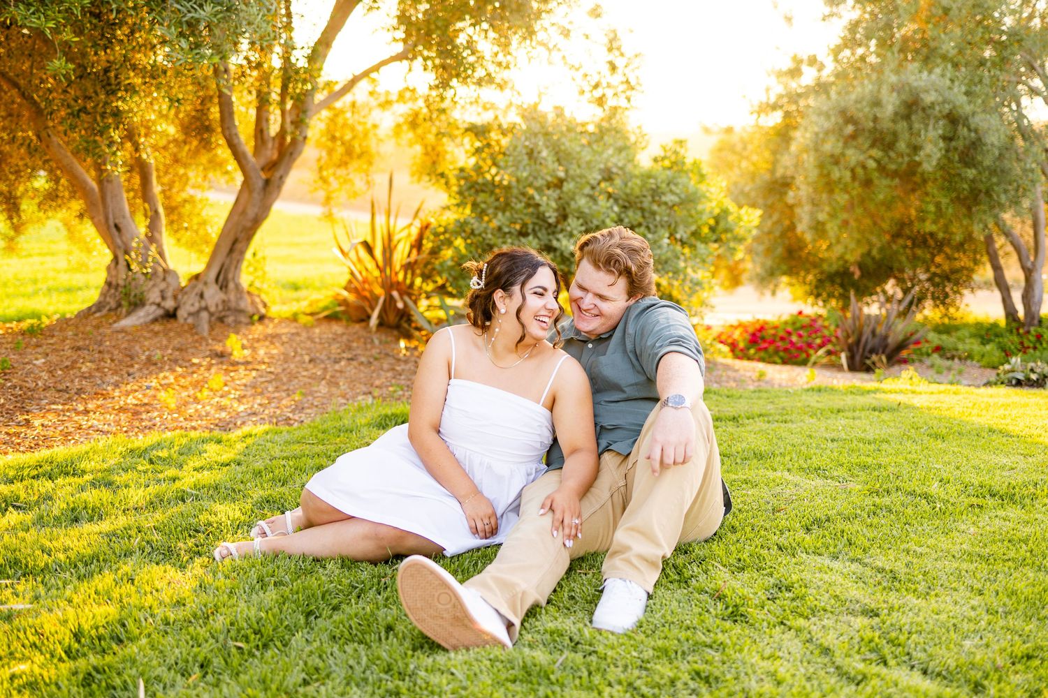 Couple relaxing on grass during golden hour with trees creating dreamy backdrop.
