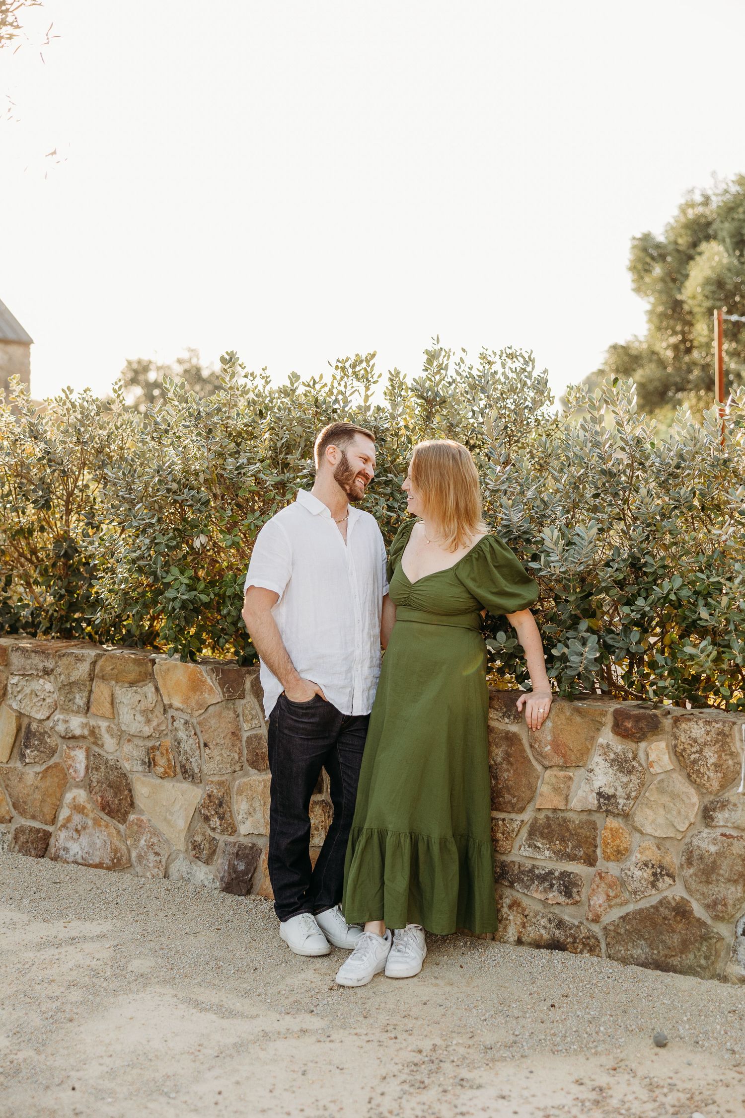 A couple sits together on a stone wall surrounded by lush greenery in matching white and olive green outfits.