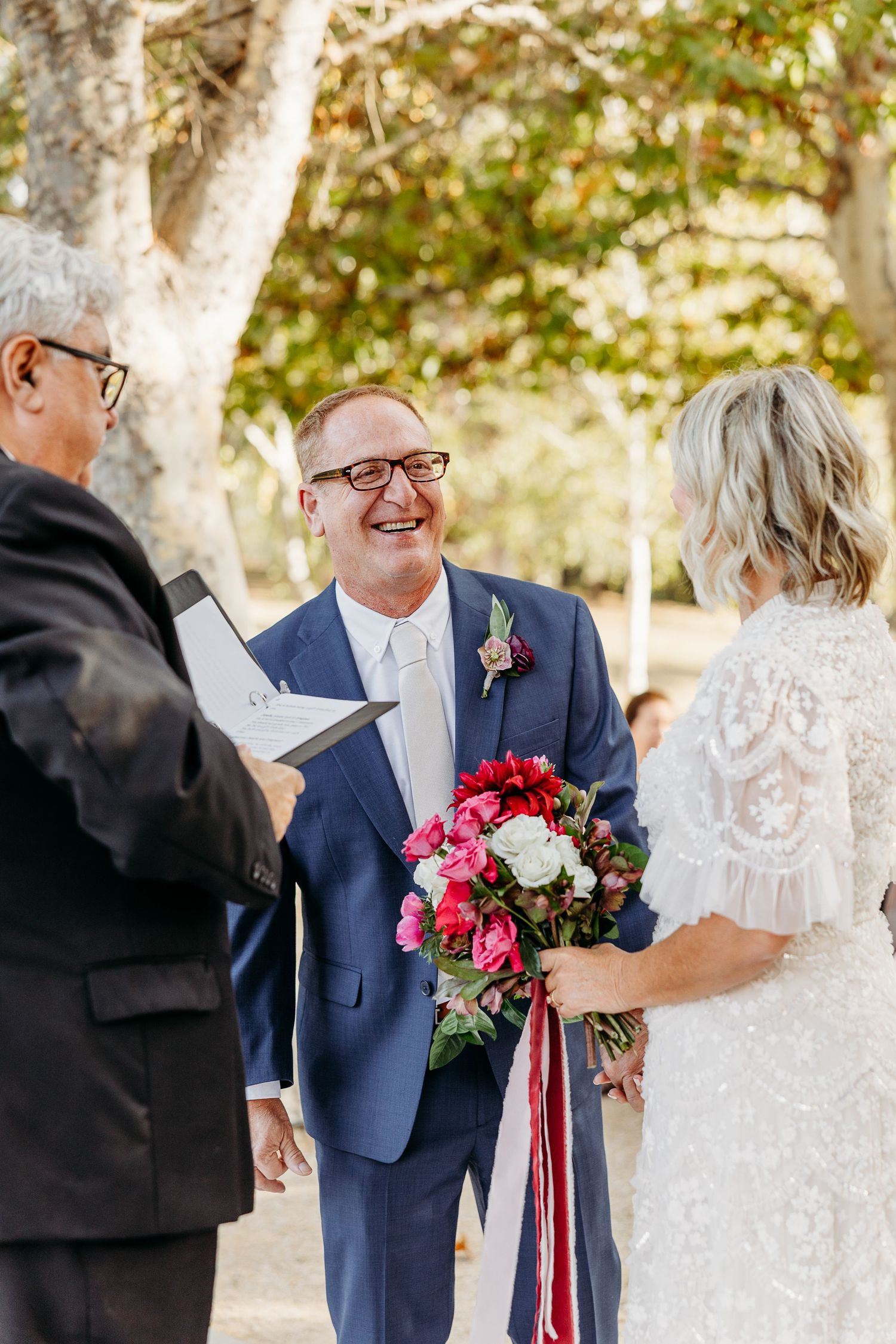 Wedding ceremony moment captured under fall trees with red and white floral bouquet.